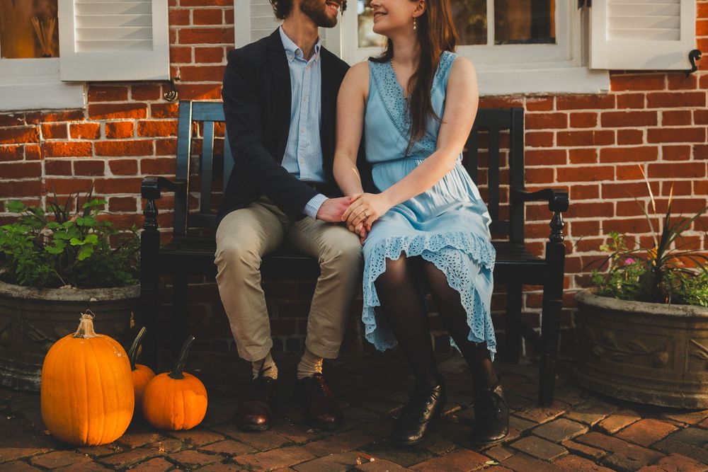 A couple sits on brick steps with autumn pumpkins beside them, wearing casual denim and blue clothing.