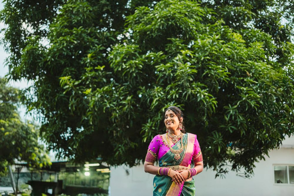 A person in a vibrant pink and green traditional silk saree stands under a lush green tree on a sunny day.