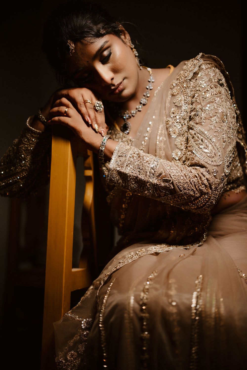 Artistic portrait in moody lighting shows a model in an ornate beige gown with intricate beadwork and embellishments.