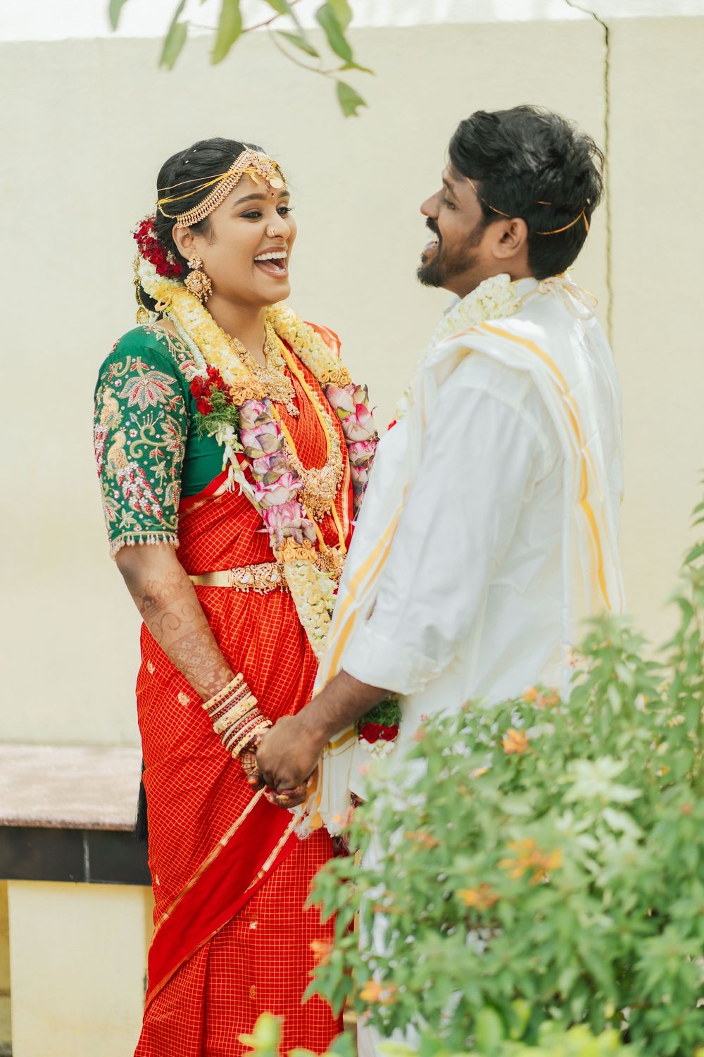 Traditional South Indian wedding couple in red and white attire pose together in garden setting.