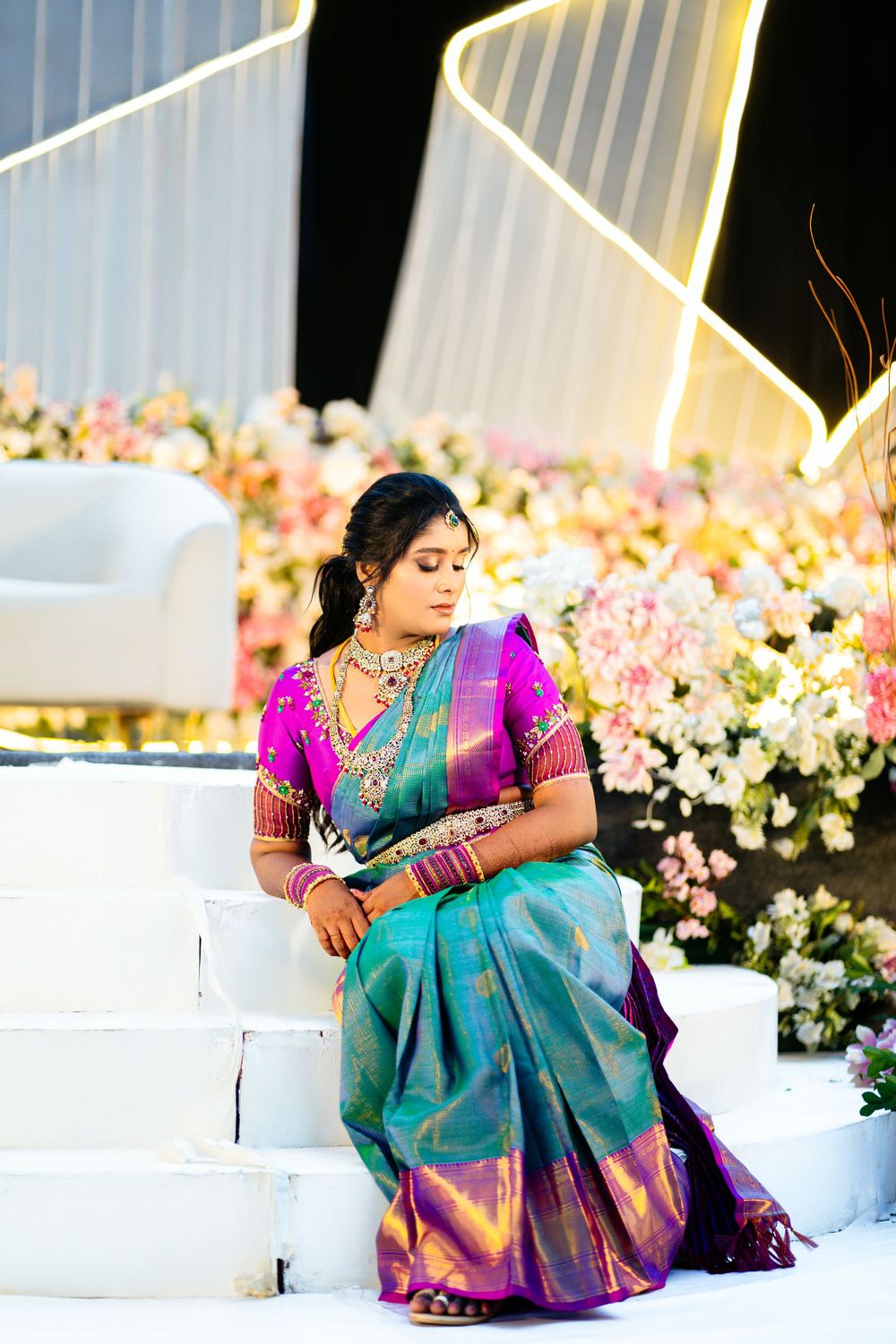 Traditional South Indian bride in turquoise and purple silk saree poses elegantly against floral backdrop.