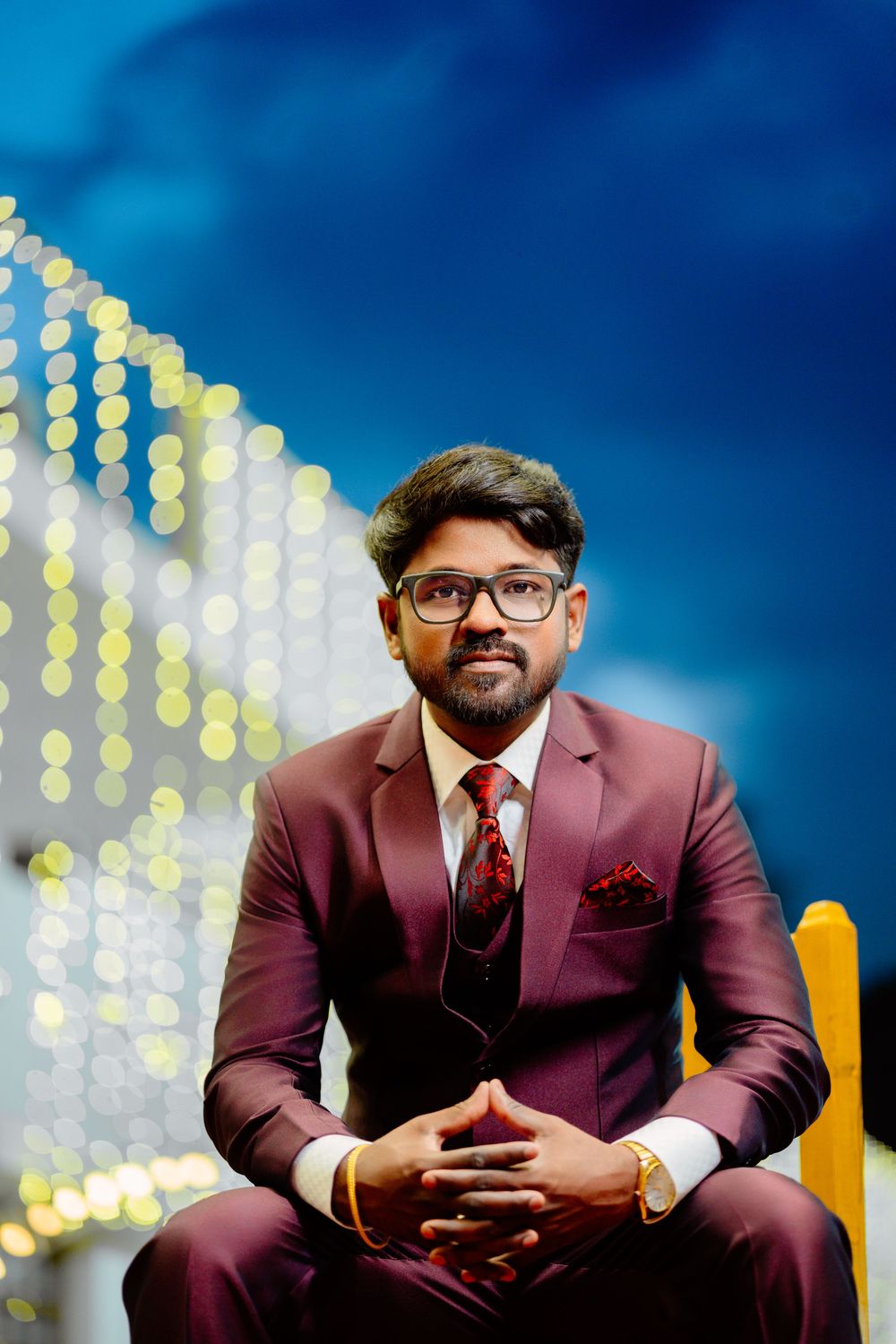 Professional portrait of someone in a burgundy suit and tie sitting against a blue background with bokeh lights.