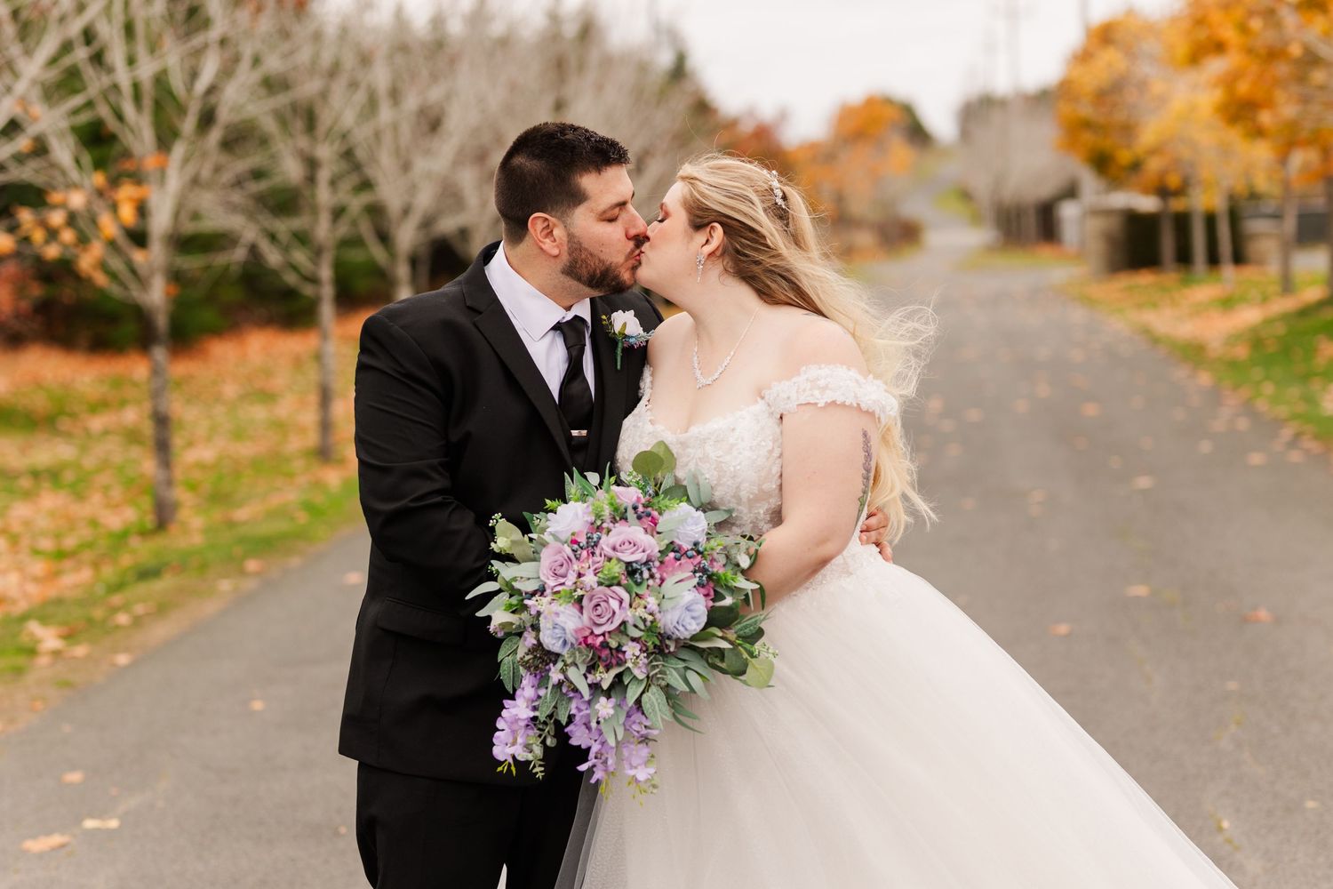 A couple in wedding attire shares an intimate moment on a tree-lined autumn road with fallen leaves.