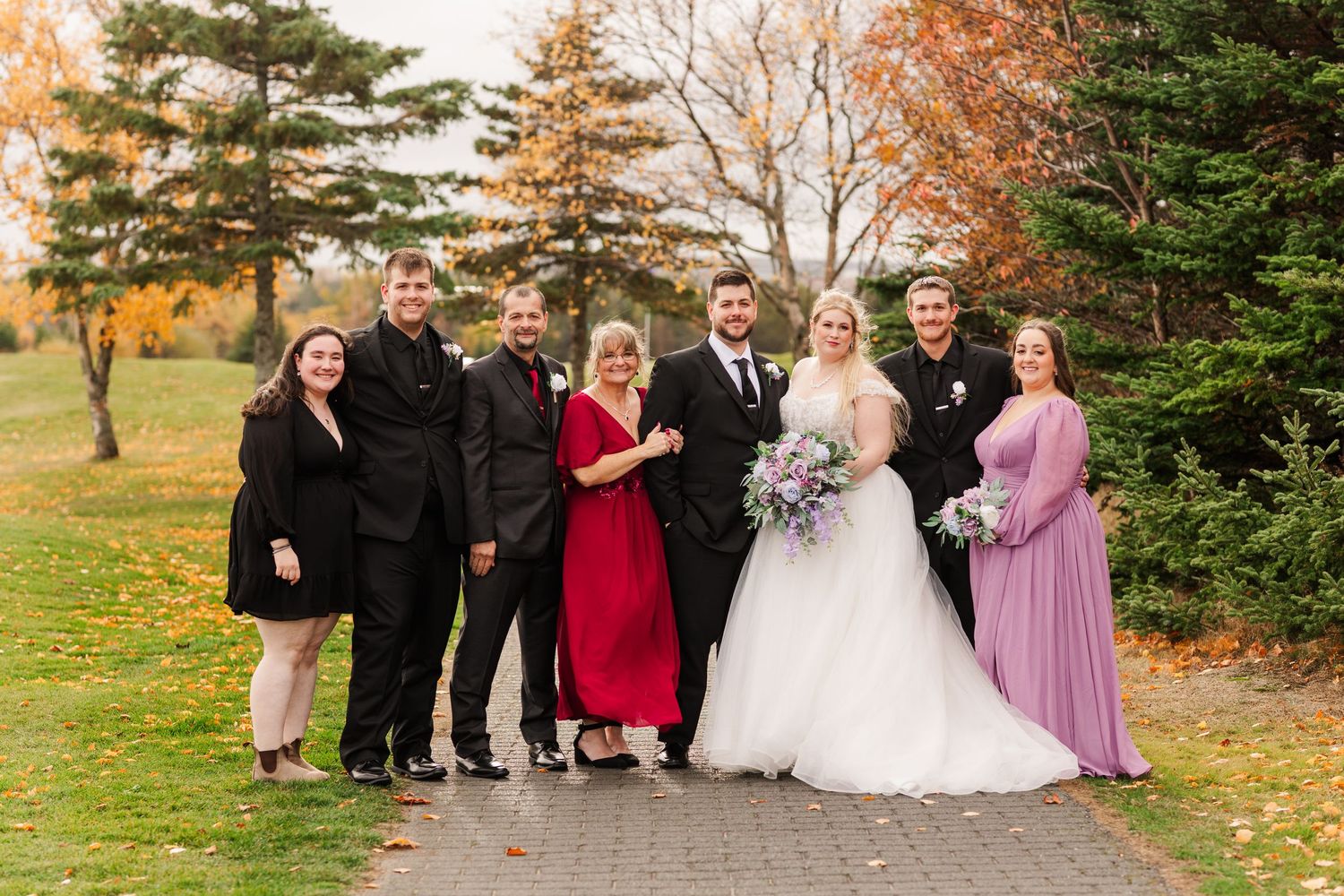 A group of wedding guests and wedding party members pose together outdoors on a fall day with autumn foliage in the background.