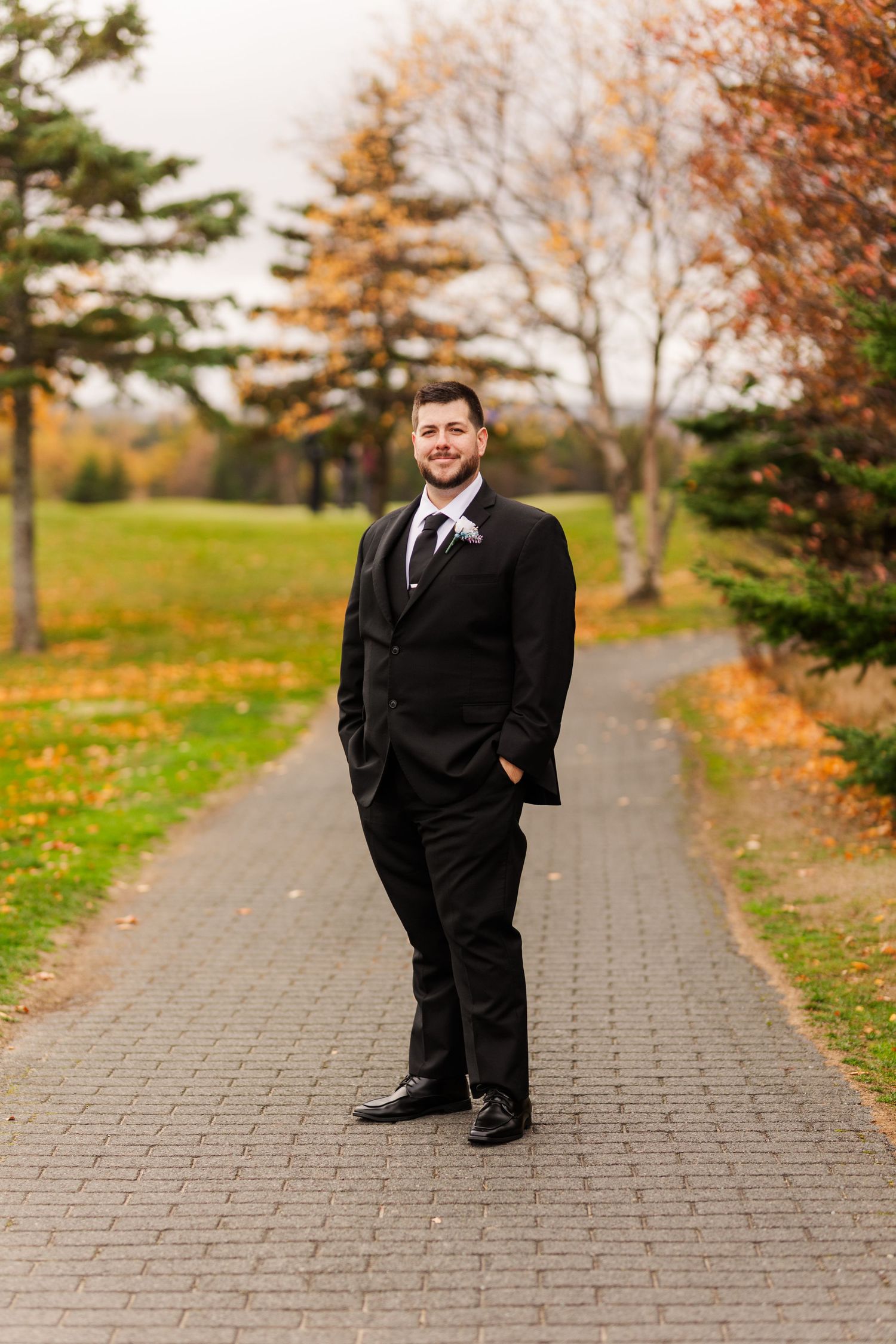 A person in a black suit stands on a stone path surrounded by autumn trees with orange and yellow foliage.