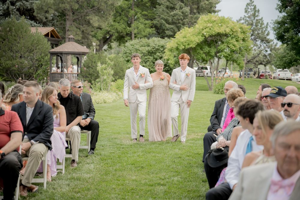 Wedding guests gather outdoors on a grassy lawn during a sunny summer ceremony in a natural garden setting.
