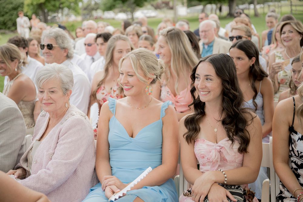 Wedding guests sit in rows of white chairs during an outdoor ceremony, wearing pastel colored dresses and smiling.