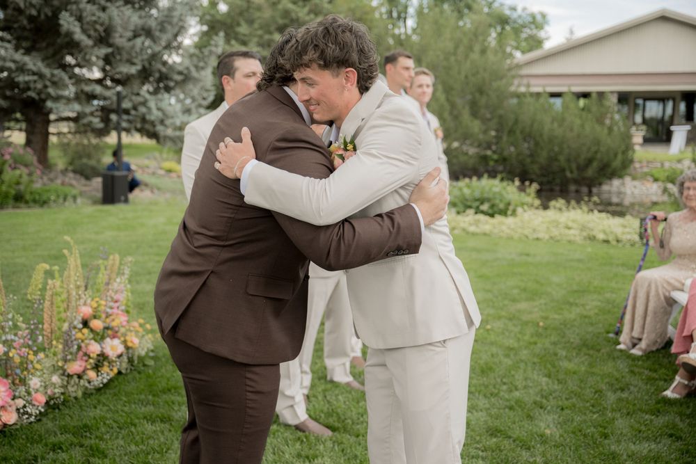 Outdoor wedding celebration showing emotional embraces between guests on a lush green lawn with trees in background.