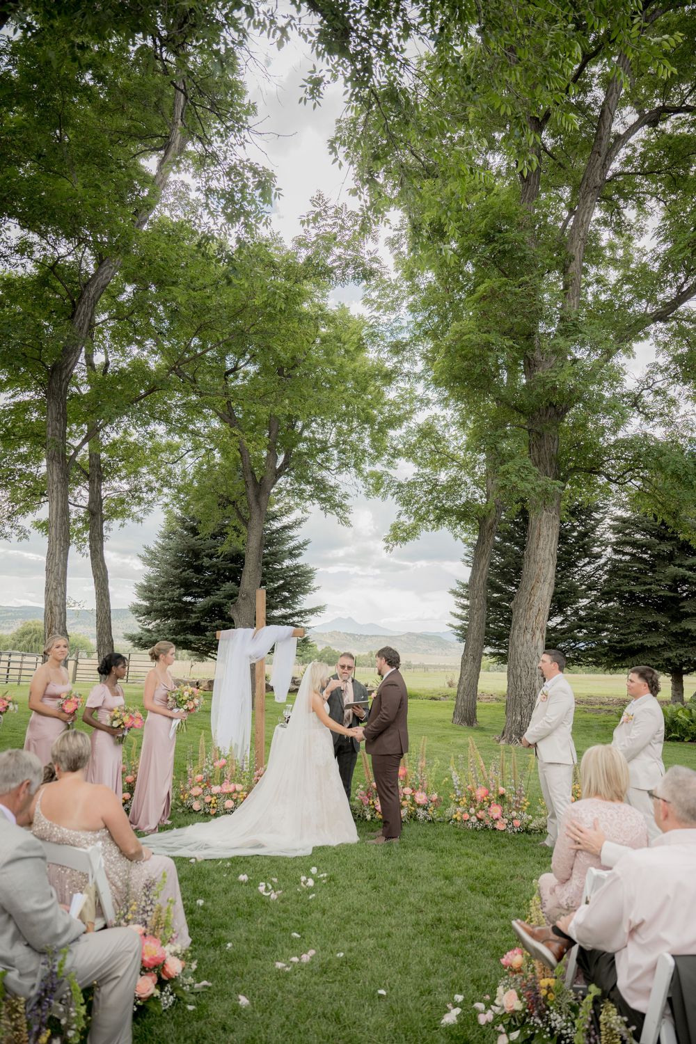 Outdoor wedding ceremony under tall trees with bridesmaids in blush pink dresses and scattered rose petals on grass.
