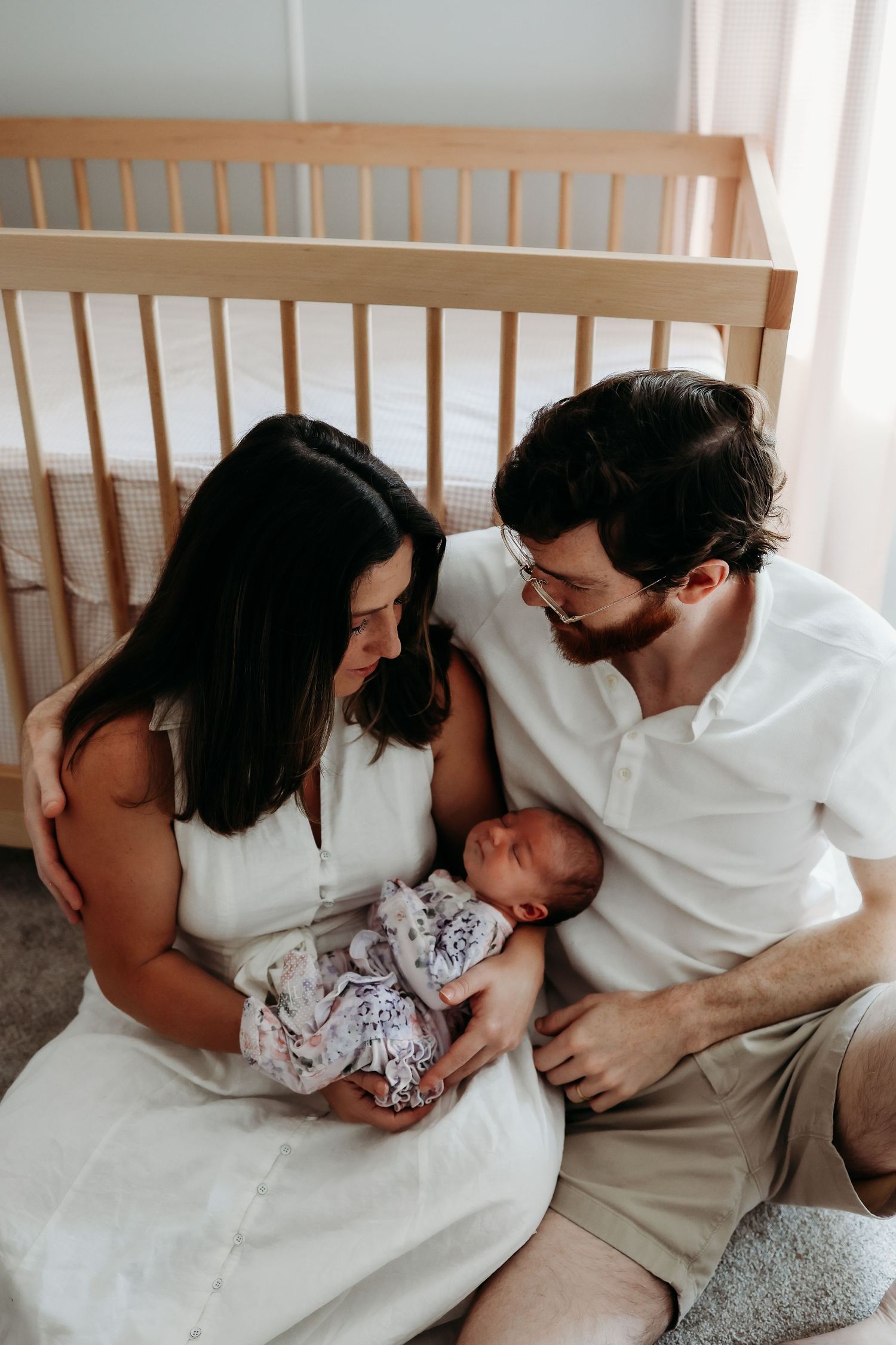 A couple sits together on a bed with their newborn baby in a nursery with a wooden crib in the background.
