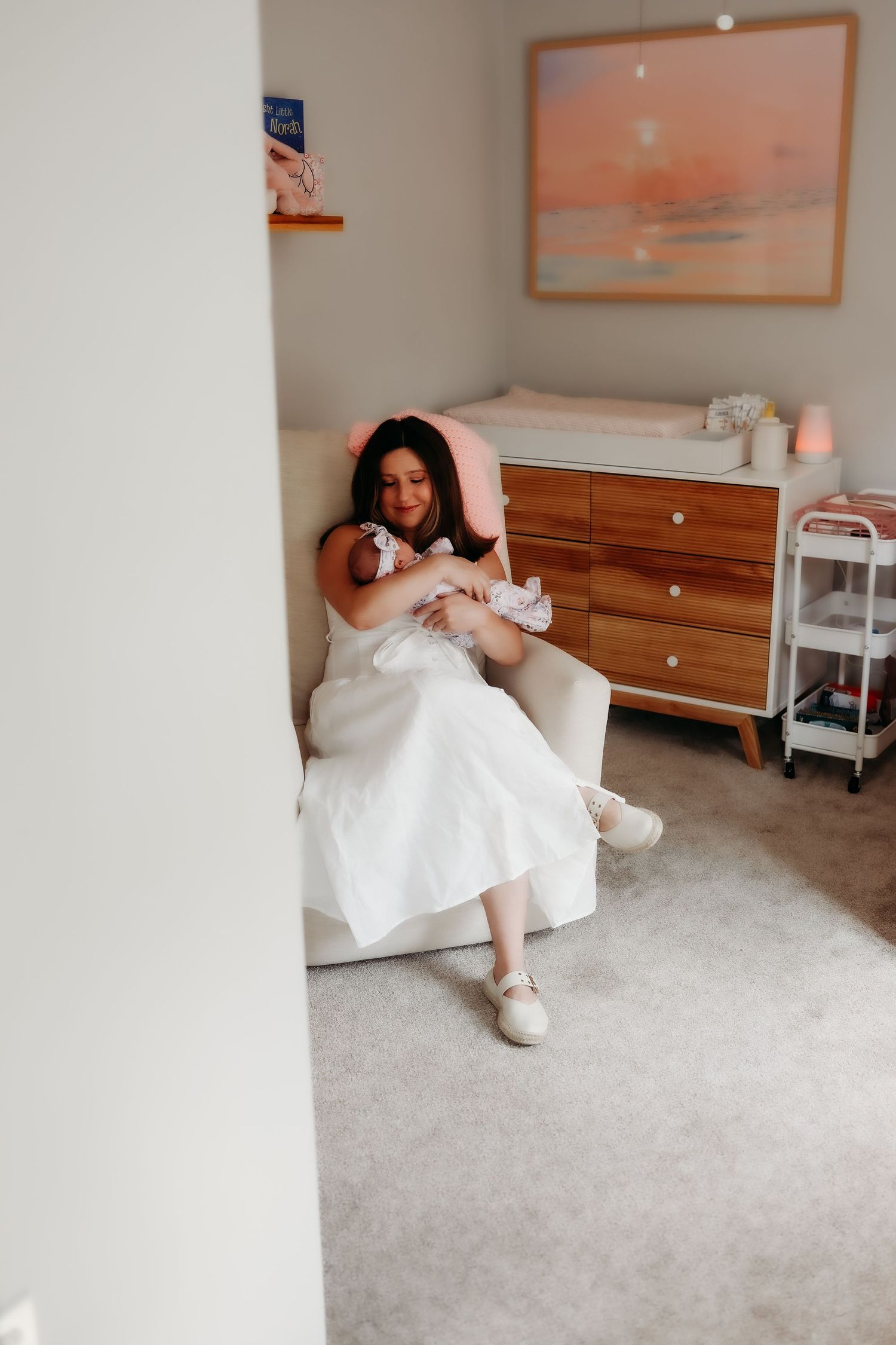 A person in a white dress sits on a carpeted floor in a minimalist room with warm-toned artwork on the walls.