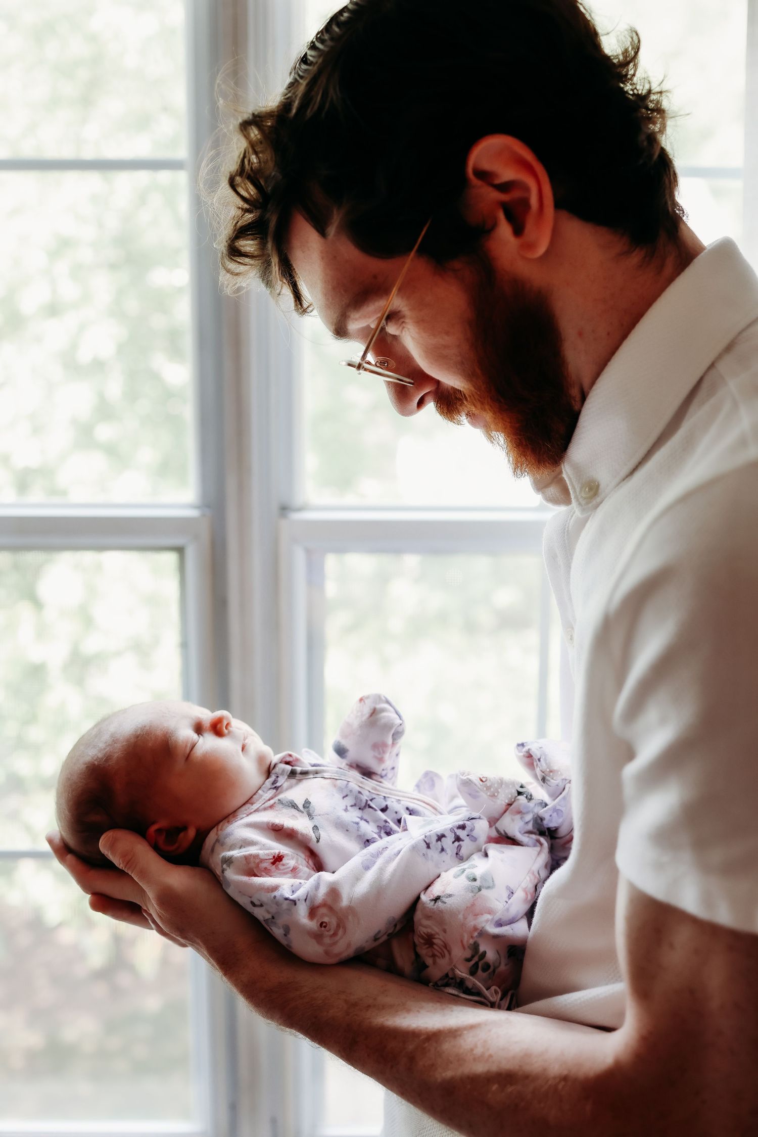 A bearded person in a white shirt holds a baby wearing floral pajamas by a bright window.