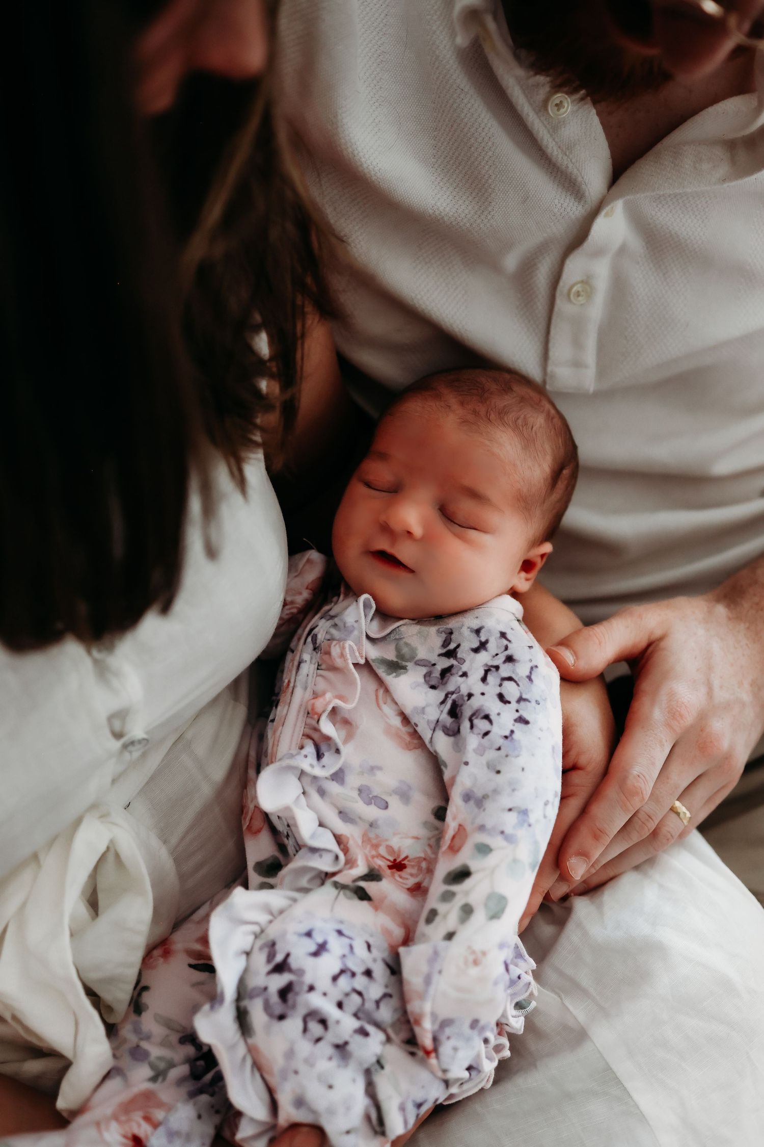 A sleeping newborn baby wearing a white patterned onesie is cradled gently in someone's arms.