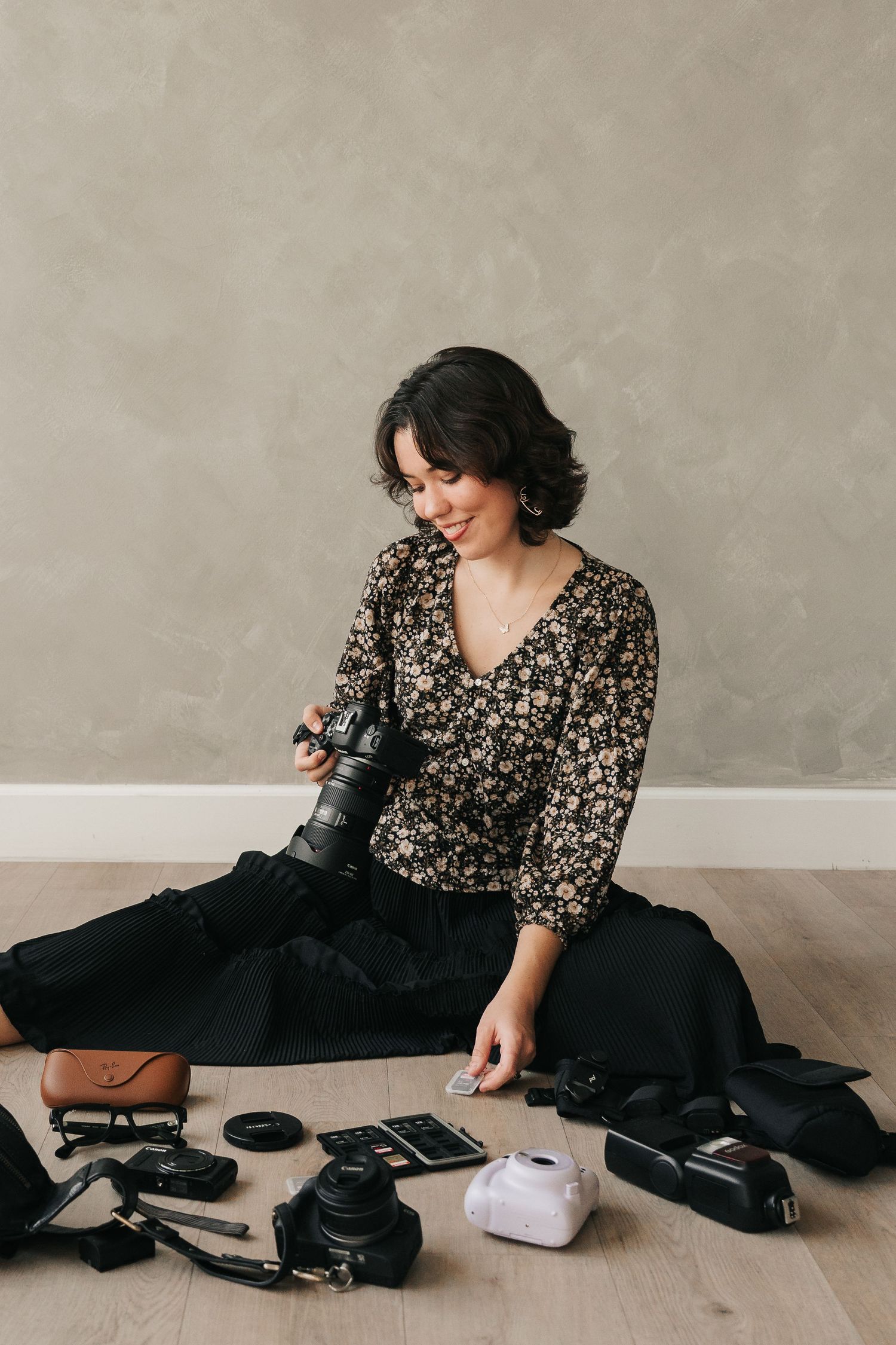 A person in a floral print top and black skirt sits on the floor surrounded by various personal items and accessories.