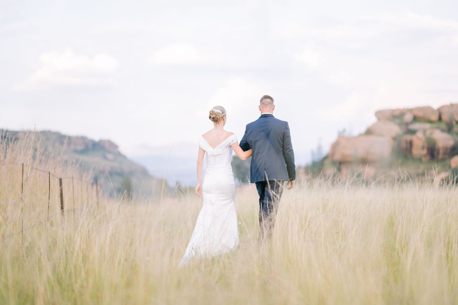 Newlyweds walk hand in hand through tall grass with mountains and rock formations in the background.