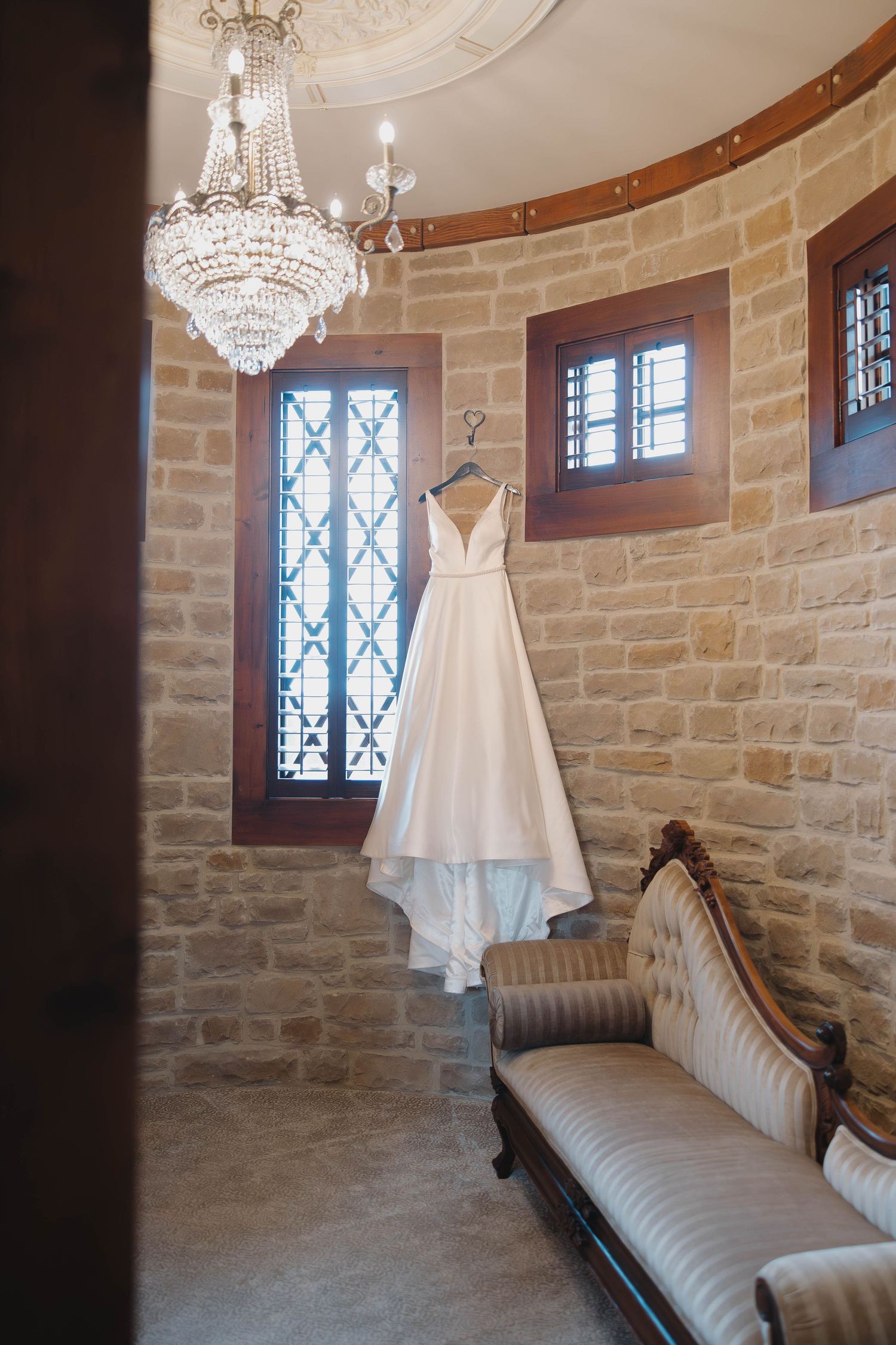 White wedding dress hangs in a rustic stone room with elegant crystal chandelier and antique chaise lounge.