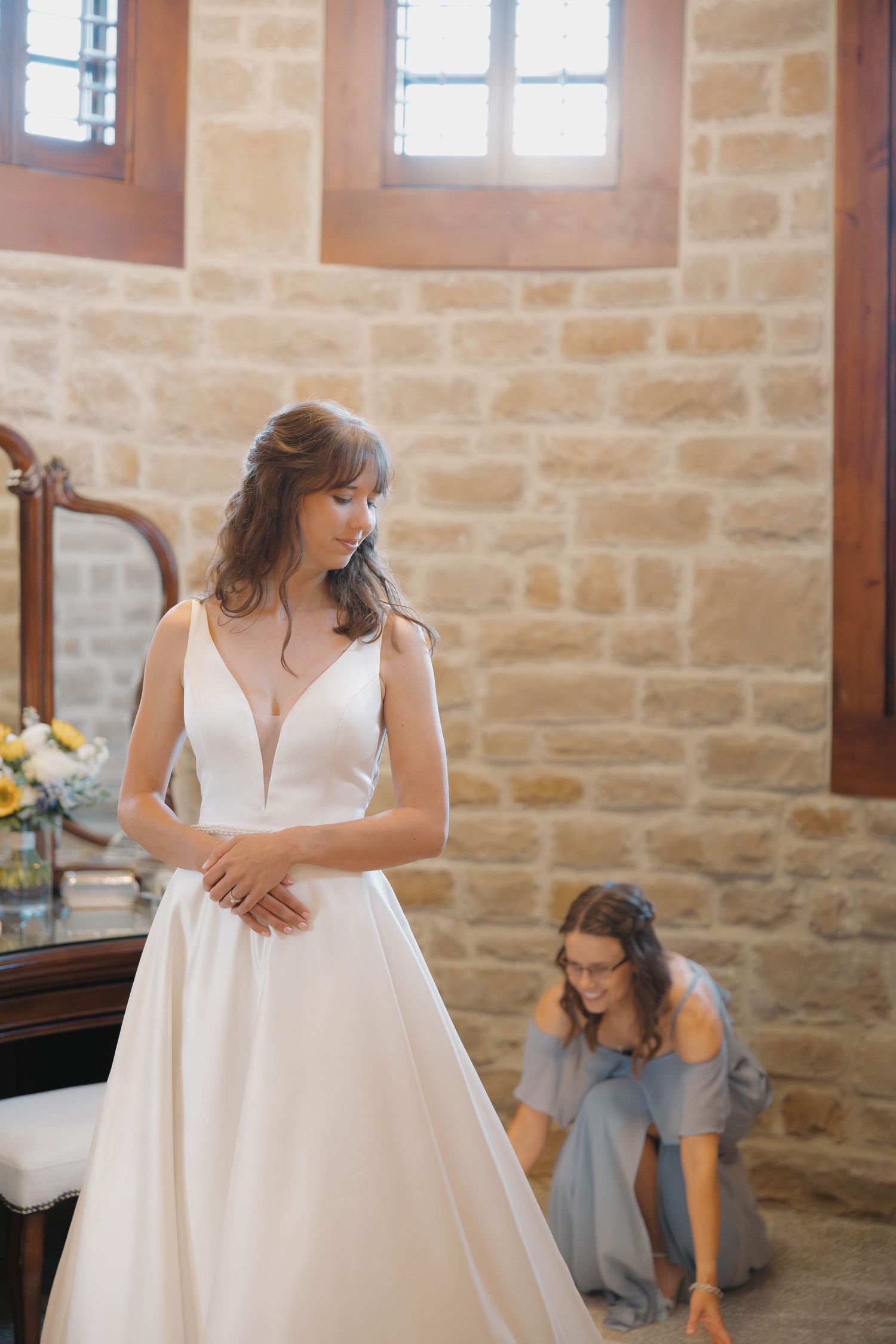 A bride in a flowing white wedding gown stands in a rustic stone-walled room with sunlit windows.