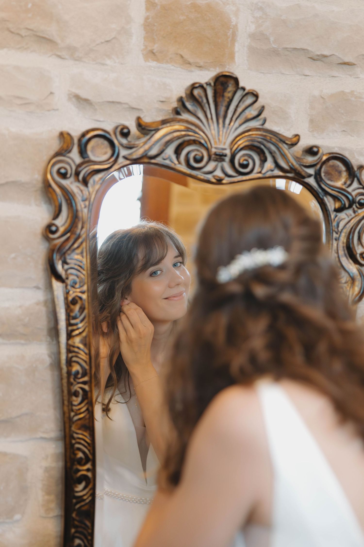A bride looks into an ornate vintage mirror during wedding preparations in a softly lit room.