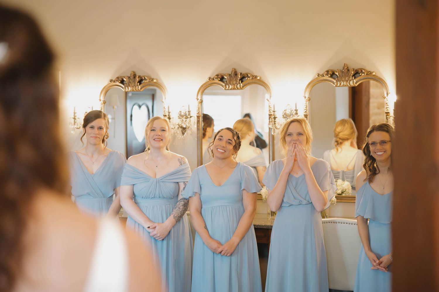 Bridesmaids in light blue dresses prepare for a wedding in an elegant room with ornate mirrors and warm lighting.