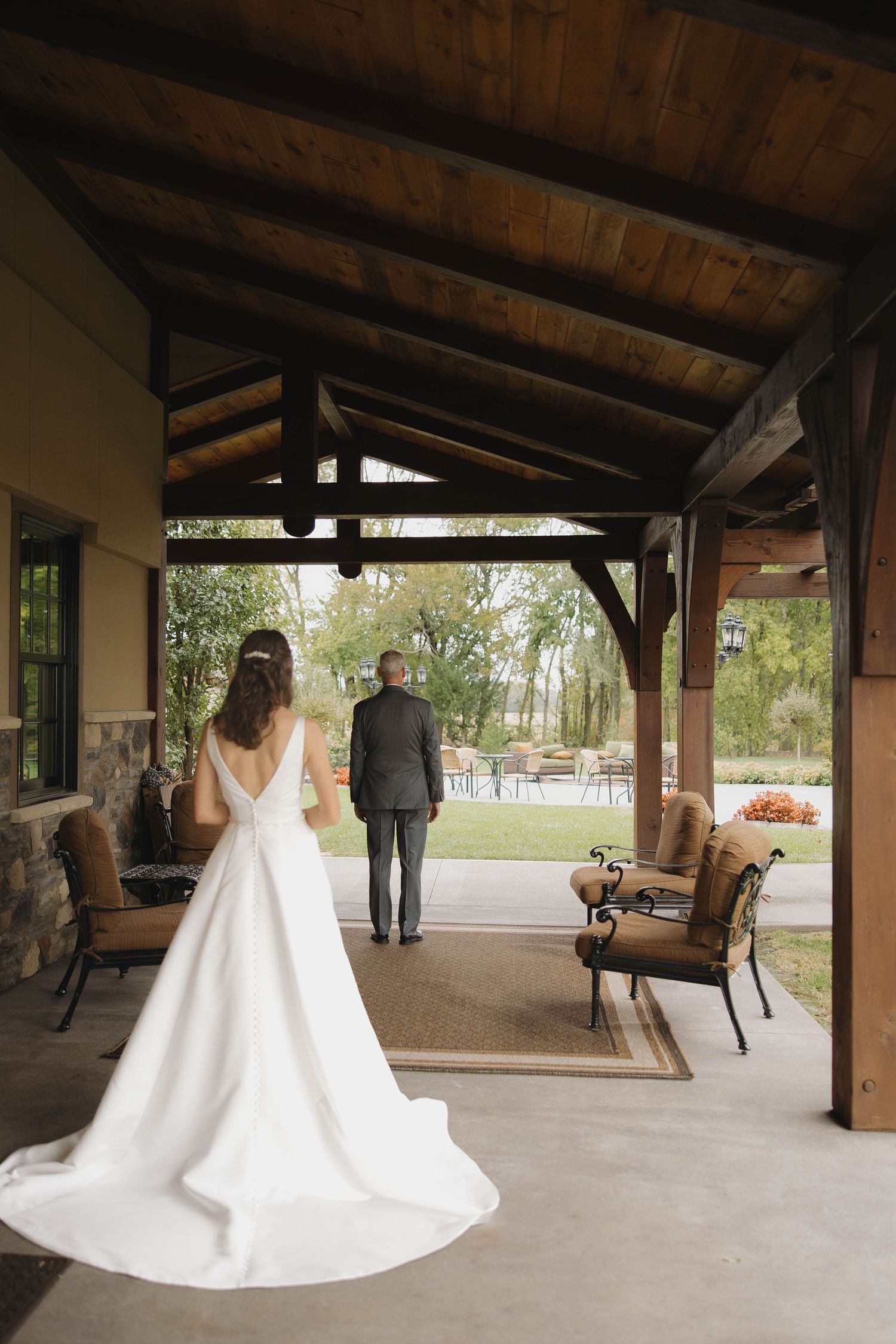 Bride in flowing white wedding dress walks along rustic covered porch as groom waits in the distance.
