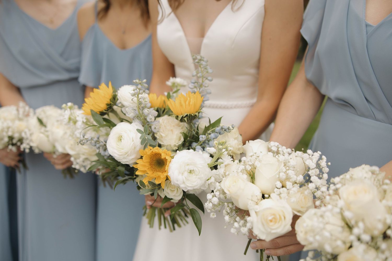 Bridesmaids in soft blue dresses hold elegant bouquets of white and yellow flowers with greenery accents.