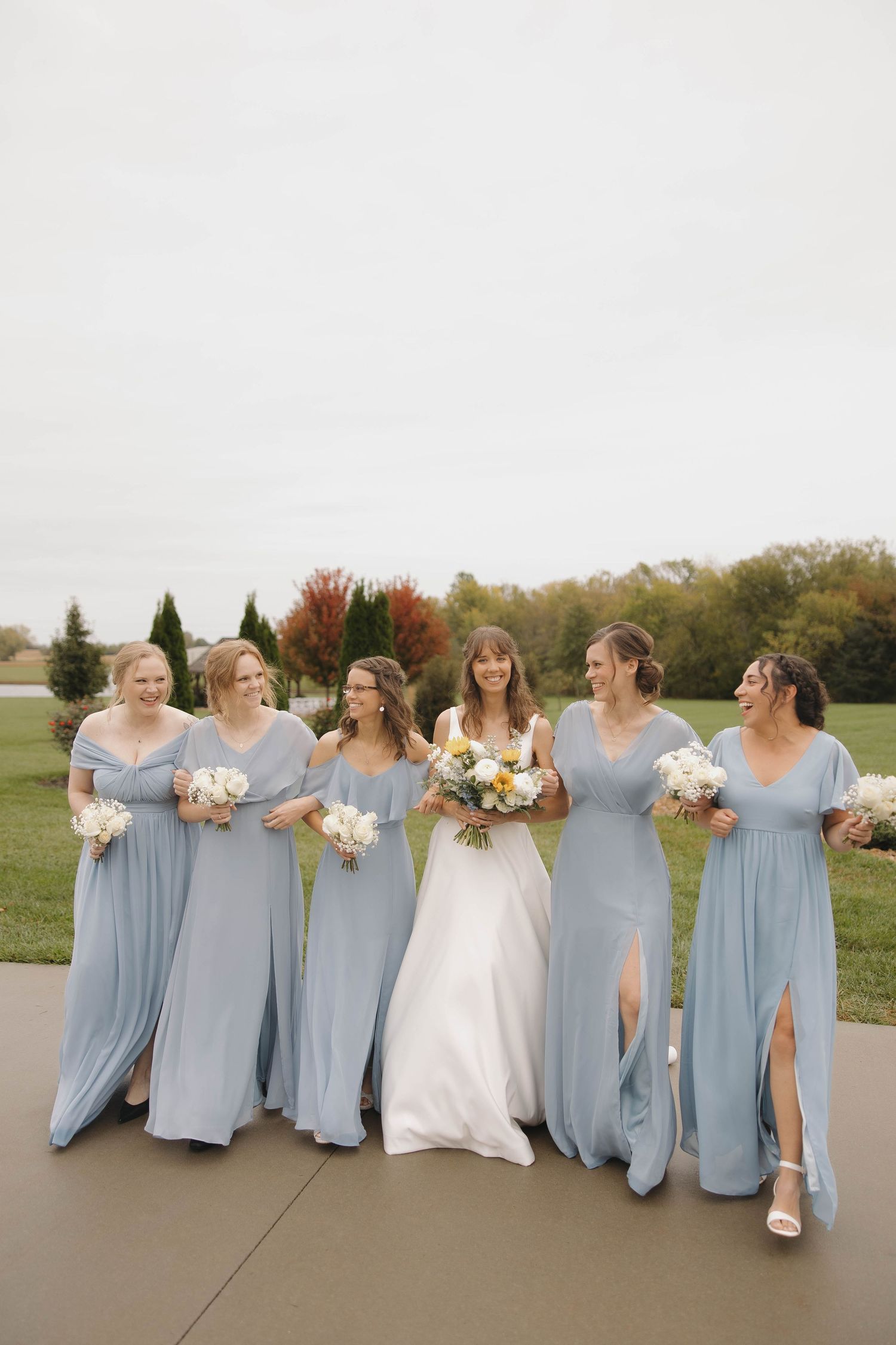 A bridal party in light blue bridesmaid dresses and white wedding gown pose together on a fall day outdoors.