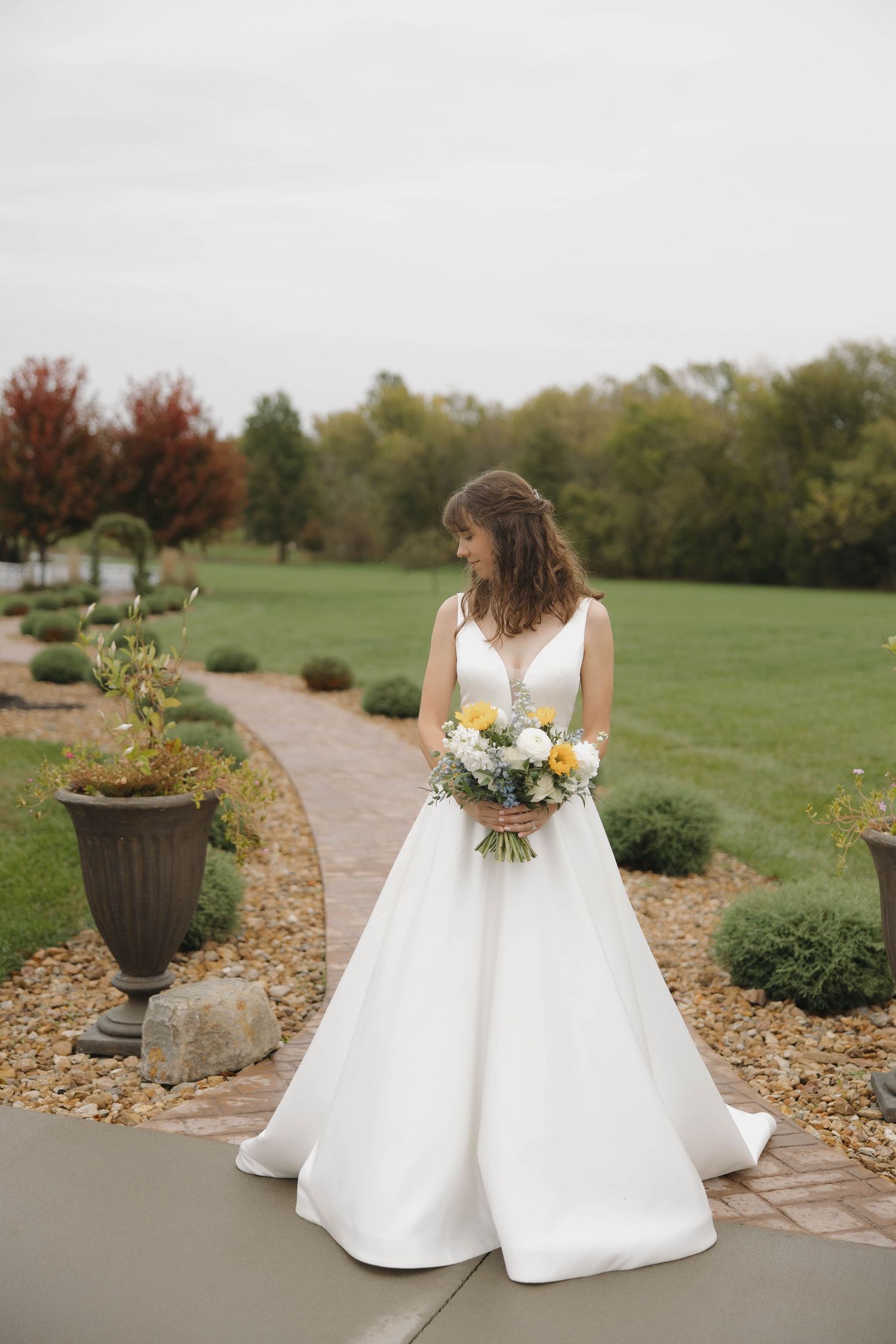 A white wedding dress with a full skirt is shown from behind on a stone path surrounded by landscaped gardens.