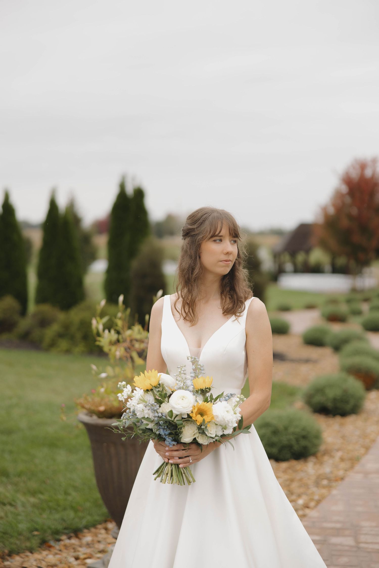 A wedding bouquet with white and yellow flowers is held against a white dress in an outdoor garden setting.