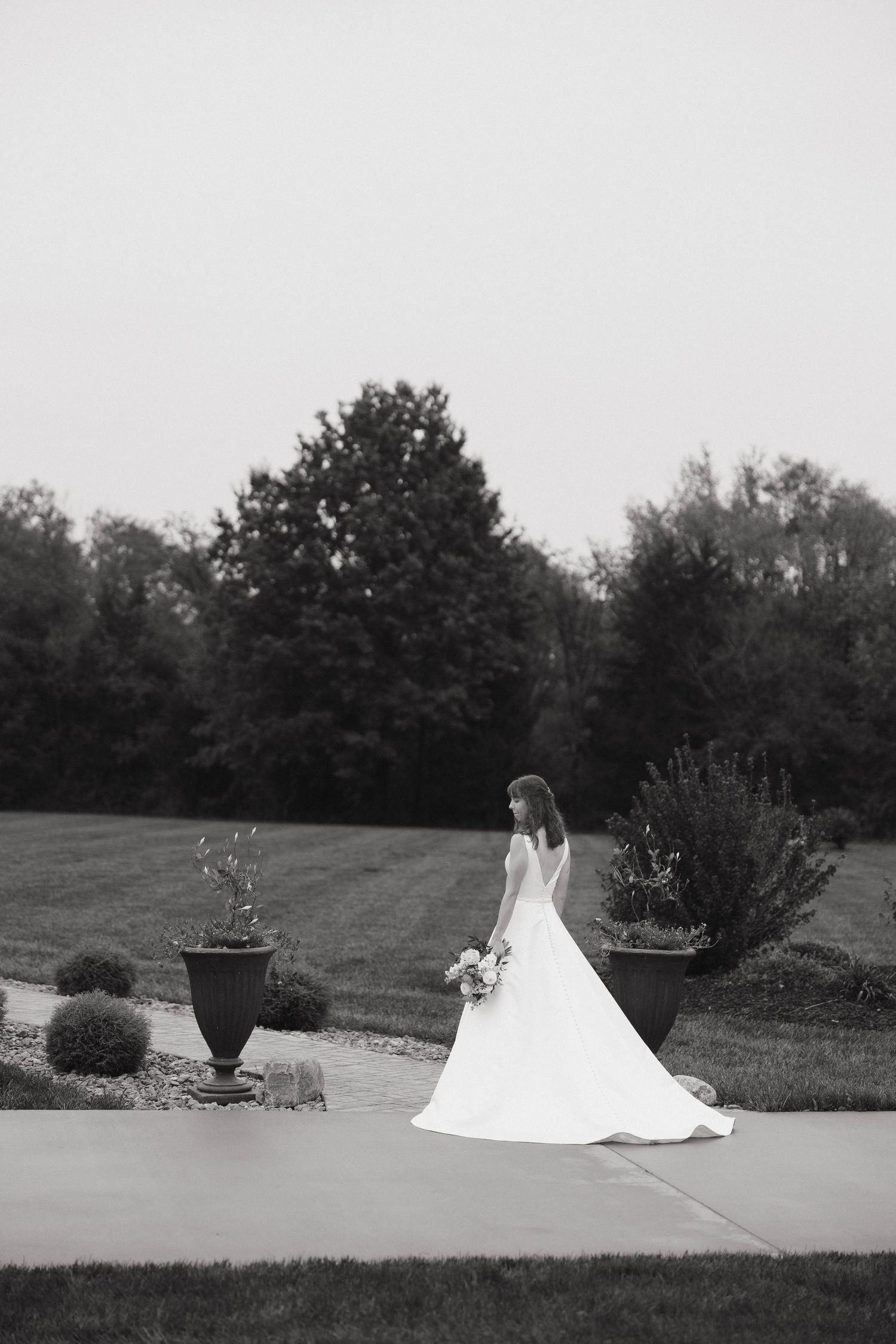 A bride in a flowing white wedding dress walks along a garden path with decorative planters and manicured grounds.
