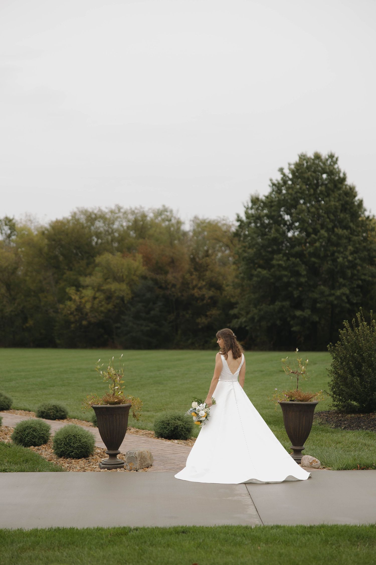 A bride in a flowing white gown sits gracefully on a stone path amidst autumn landscaping and decorative planters.