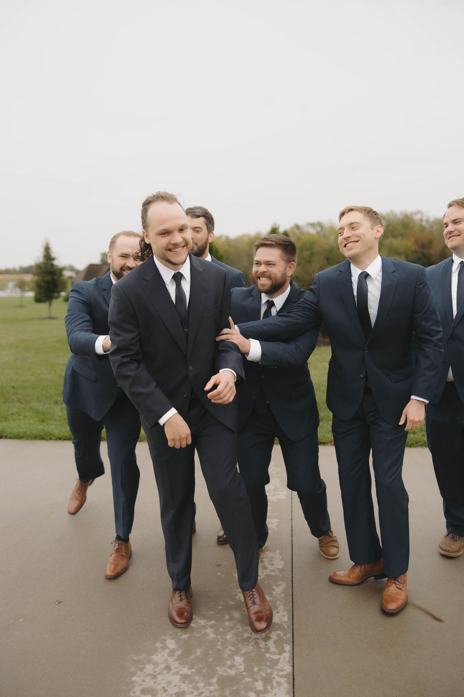 Groomsmen in navy suits and brown shoes pose playfully for a group photo on a concrete path.
