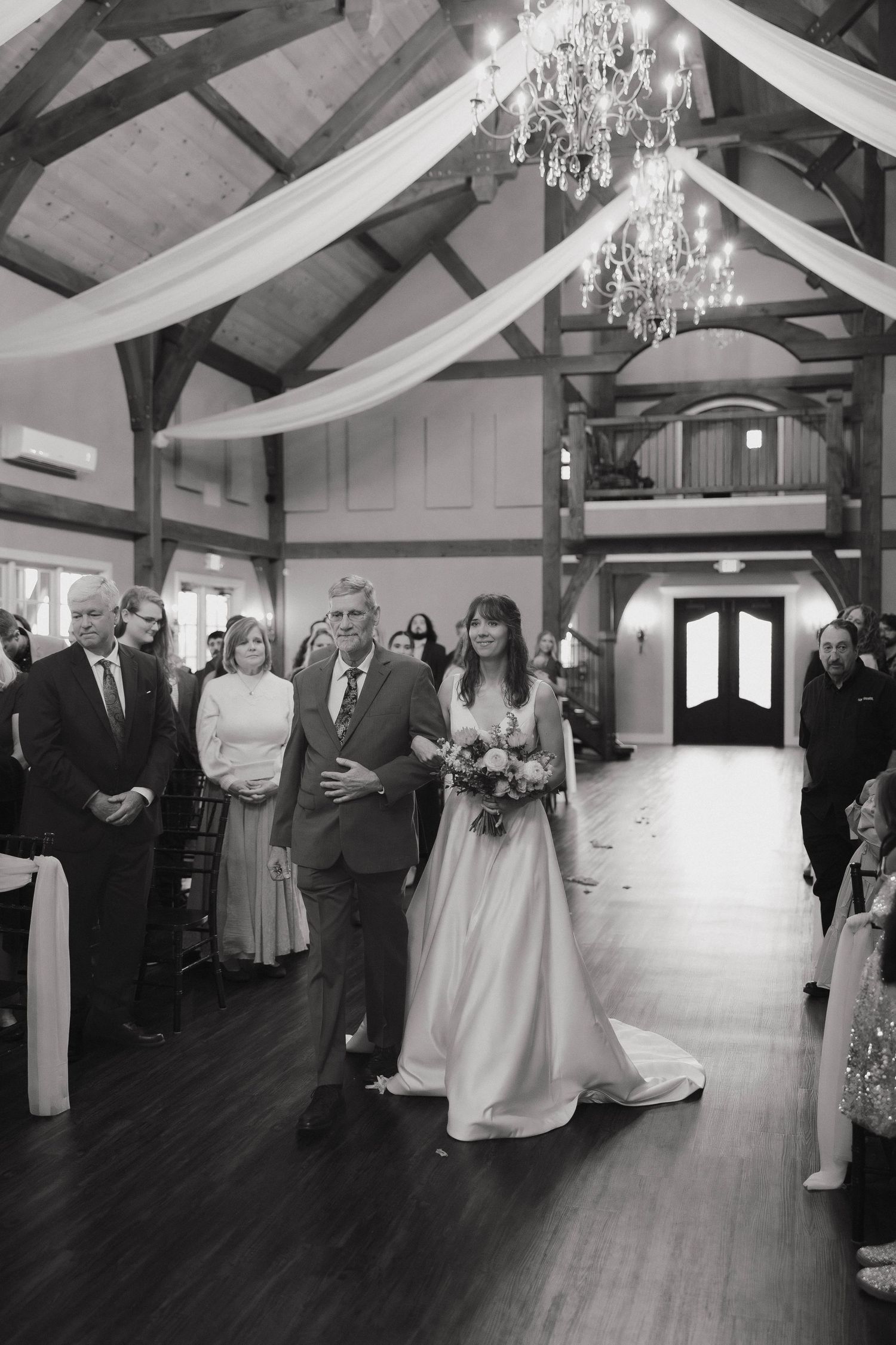 Wedding ceremony in a rustic indoor venue with white drapes and chandeliers hanging from exposed wooden beams.