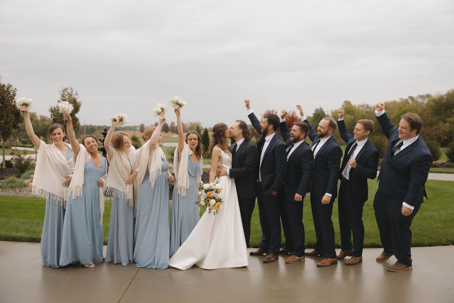 Wedding party in blue dresses and dark suits poses together on grassy hillside during sunset celebration.