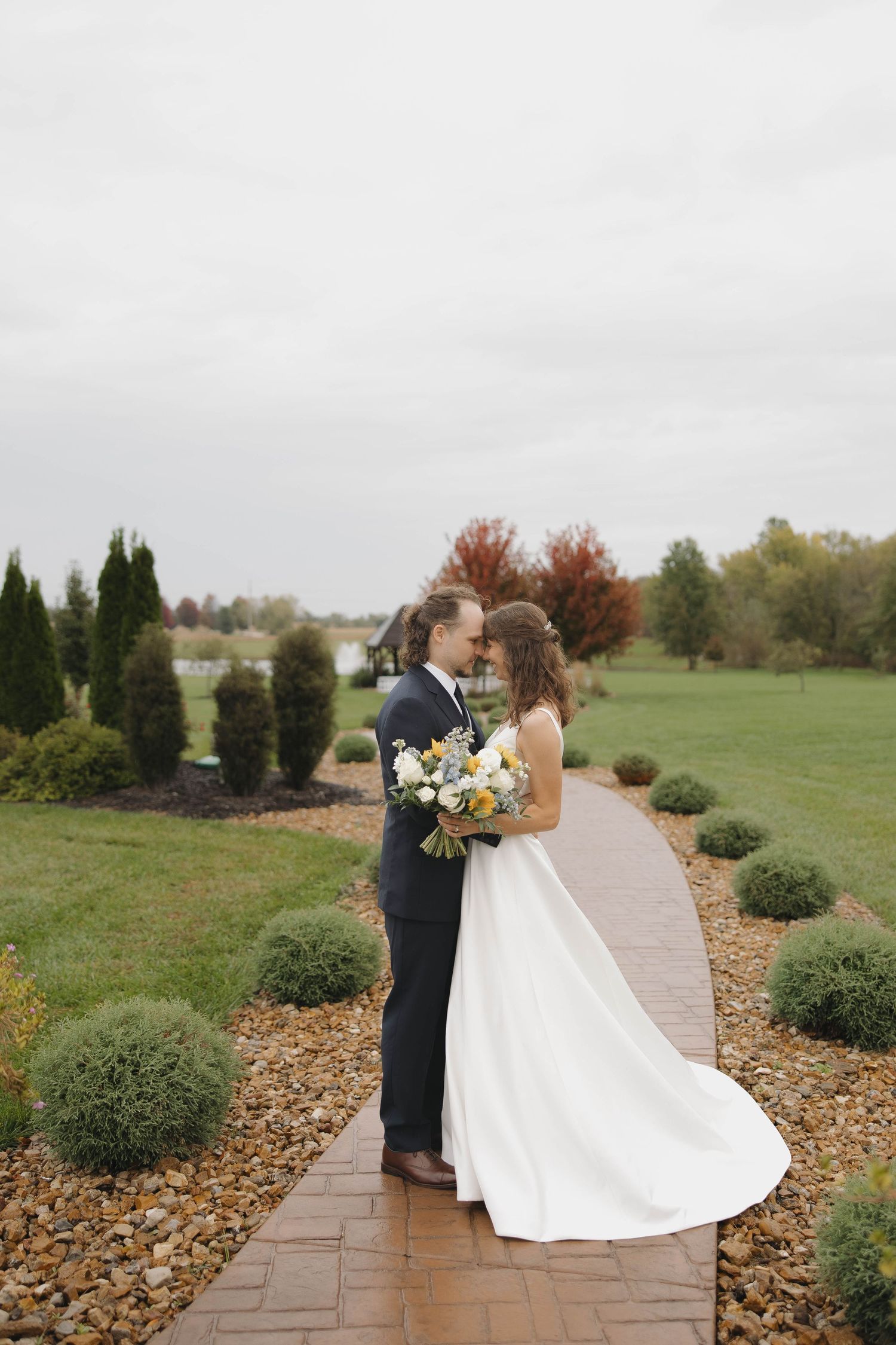 Couple in wedding attire share intimate moments along a garden path surrounded by autumn foliage and landscaping.