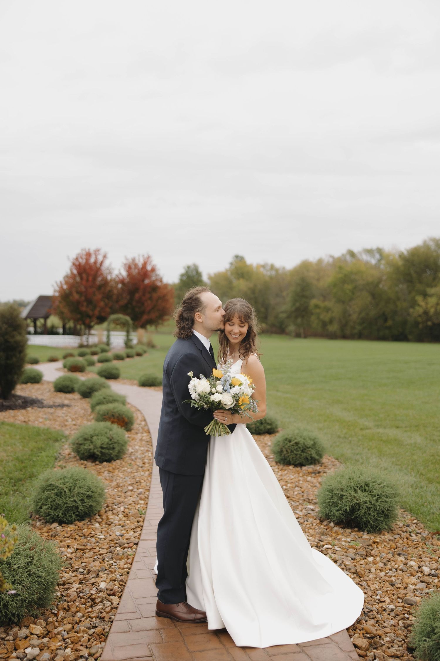 A couple shares a romantic moment in their wedding attire on a scenic path surrounded by fall foliage and green lawns.