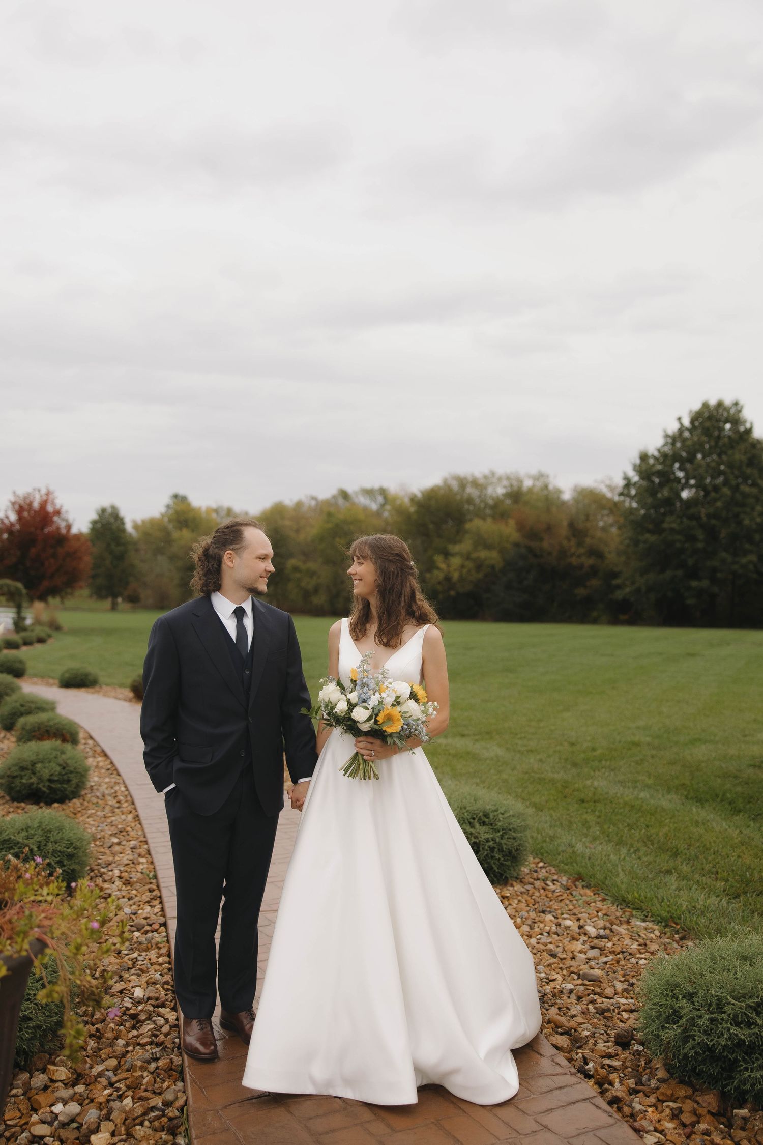 Wedding couple walking along a stone path surrounded by autumn foliage and green lawn.