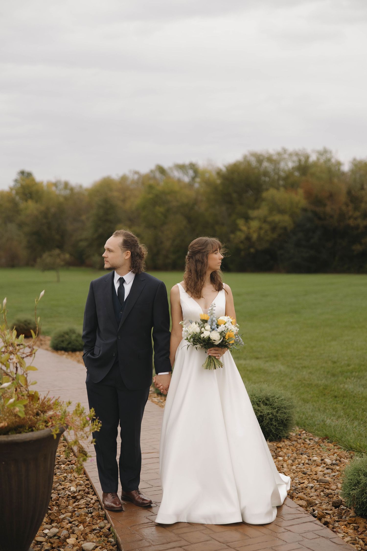 Newlyweds stroll along a landscaped pathway with fall colors and open fields in the background.