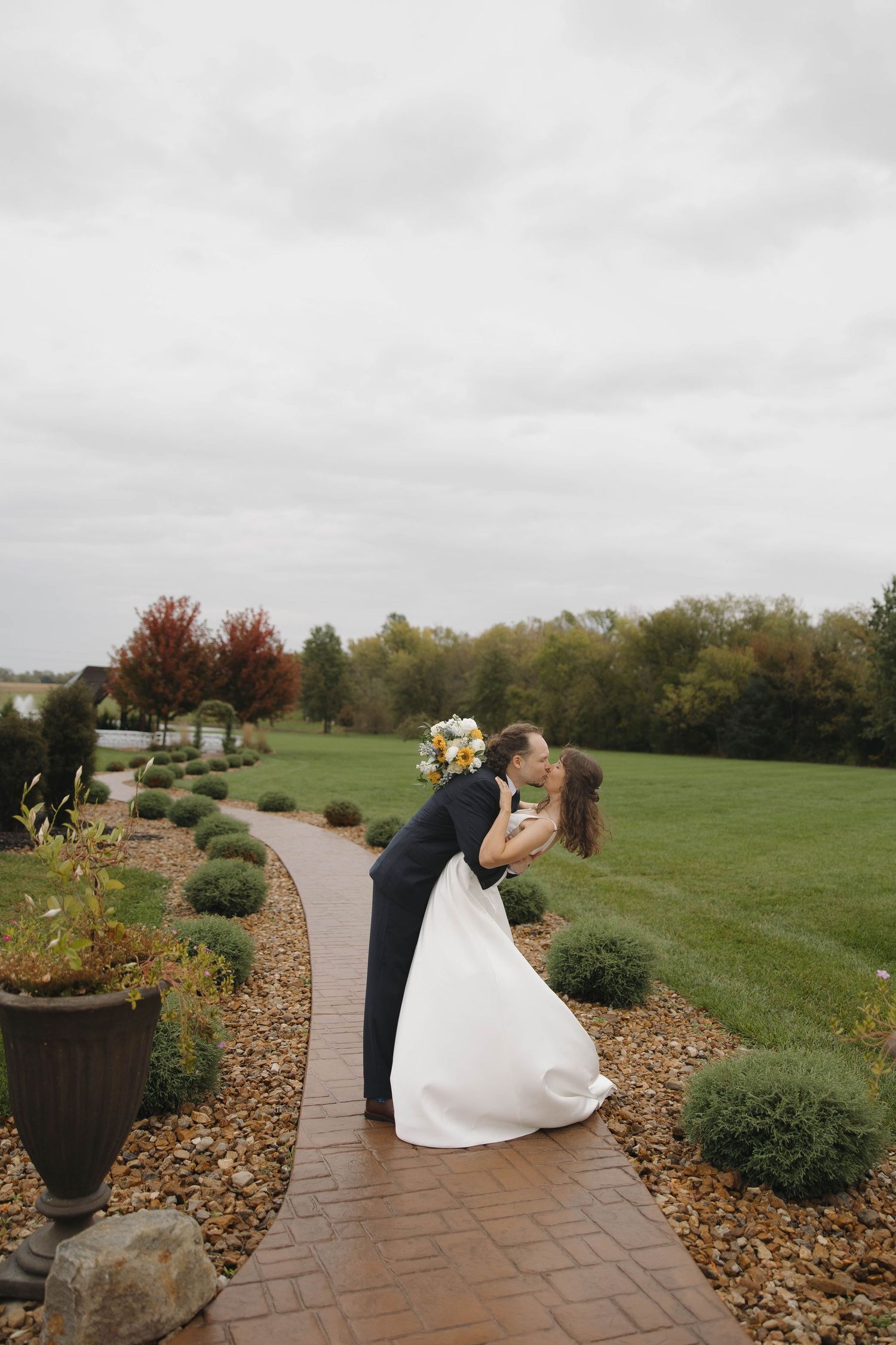 Romantic couple embraces on a winding stone path surrounded by autumn foliage and rolling hills at a scenic venue.