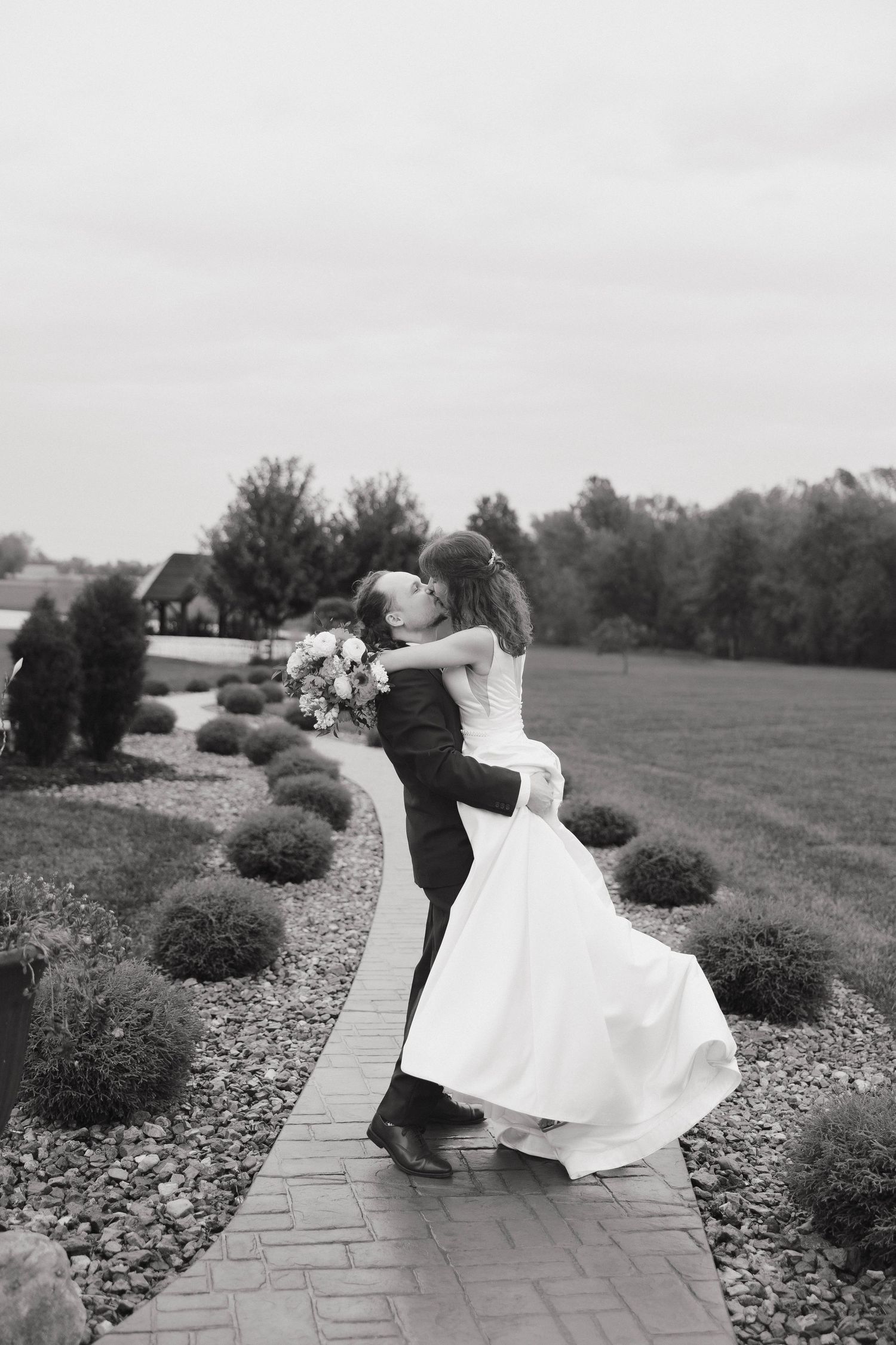 Black and white photo of a couple dancing playfully on a stone pathway surrounded by landscaping.