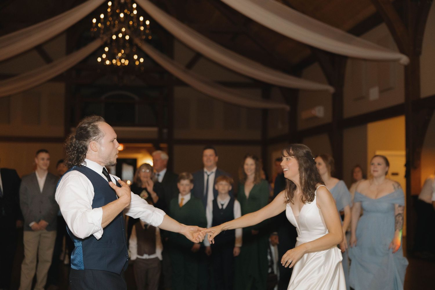 Wedding guests and bridal party dancing with arms raised in a dimly lit reception venue.