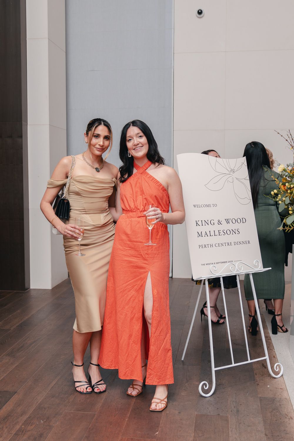 Two guests in elegant dresses pose next to a welcome sign at an event venue.