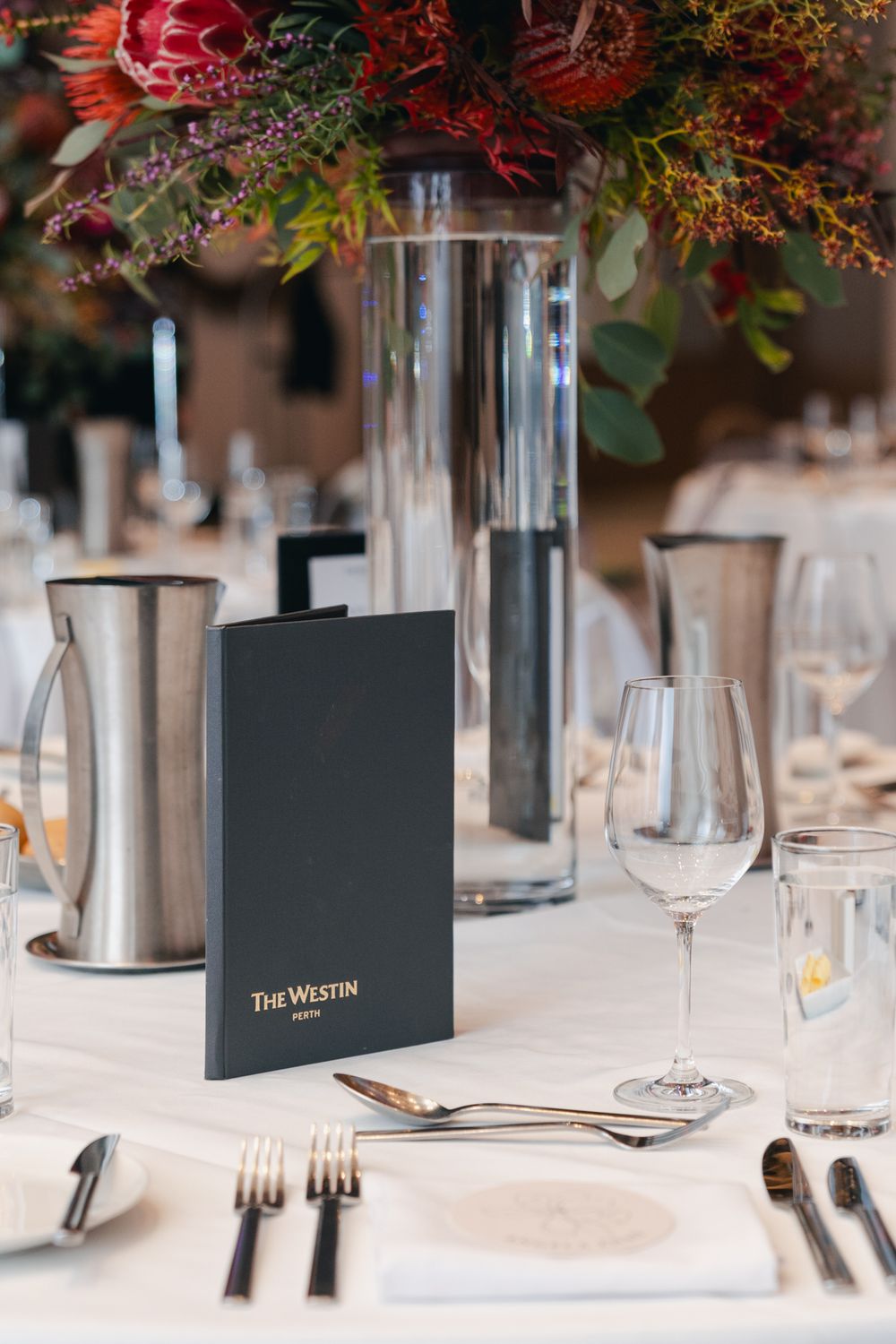 Elegant table setting with wine glasses, silverware and a Westin menu card under red floral centerpiece.
