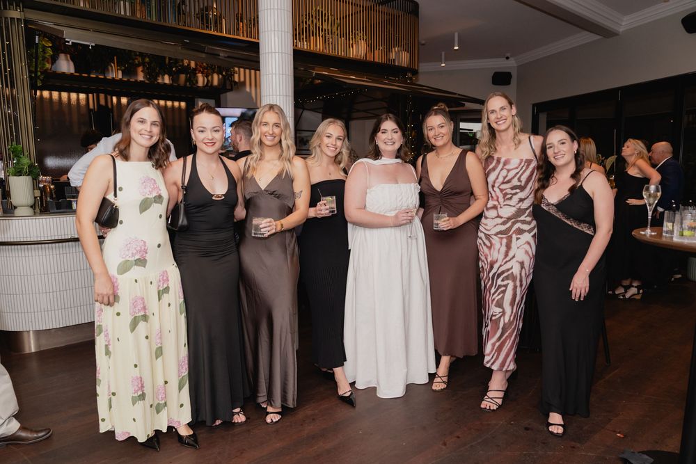 A group of people in formal dresses pose together at an elegant party venue with a bar in the background.
