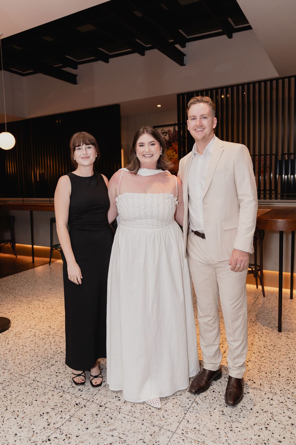 Three people pose together at an elegant event, with one wearing a black dress and two in light-colored formal attire.