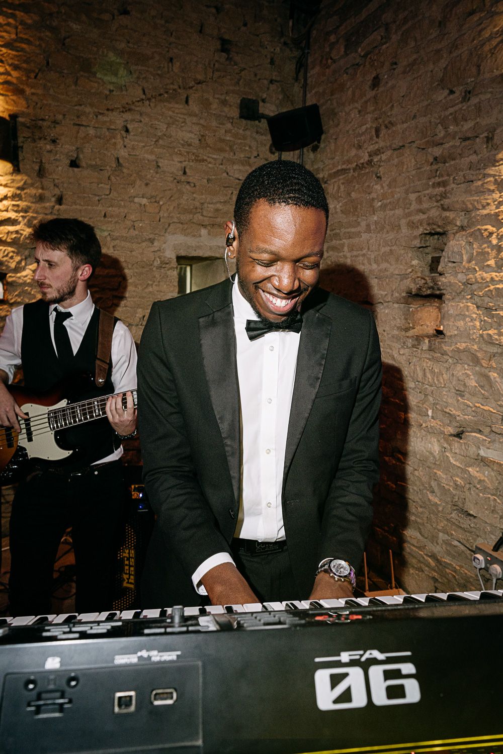 DJ in formal black tuxedo and white shirt smiles while performing at an event with brick wall backdrop.