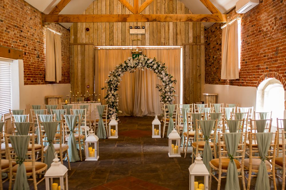 Elegant barn wedding ceremony setup with wooden walls, exposed beams, and white chairs with sage green accents.