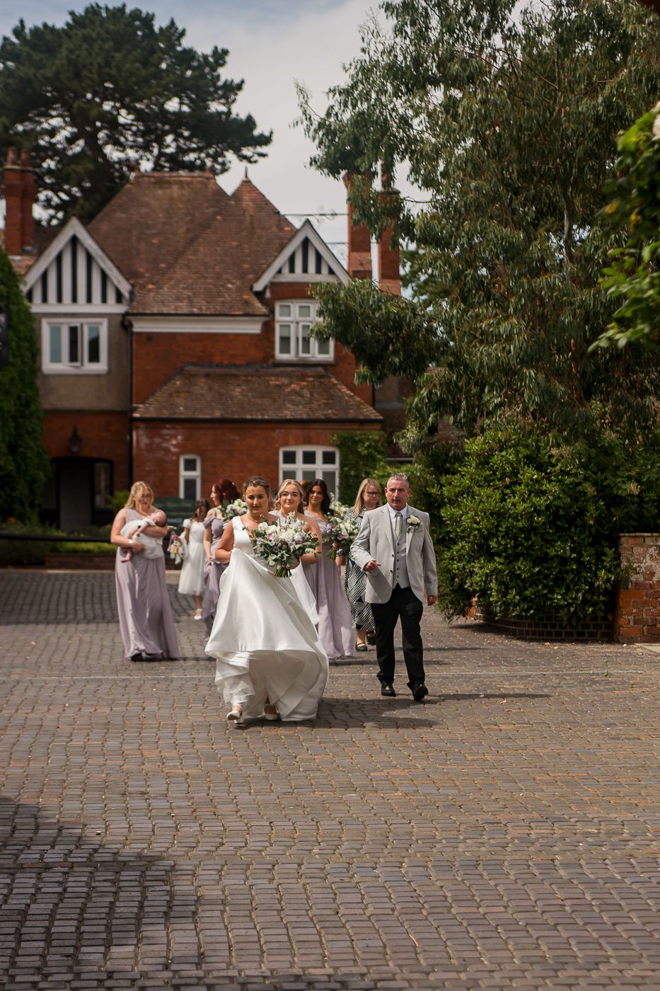 Wedding party walking across cobblestone courtyard in front of brick manor house.