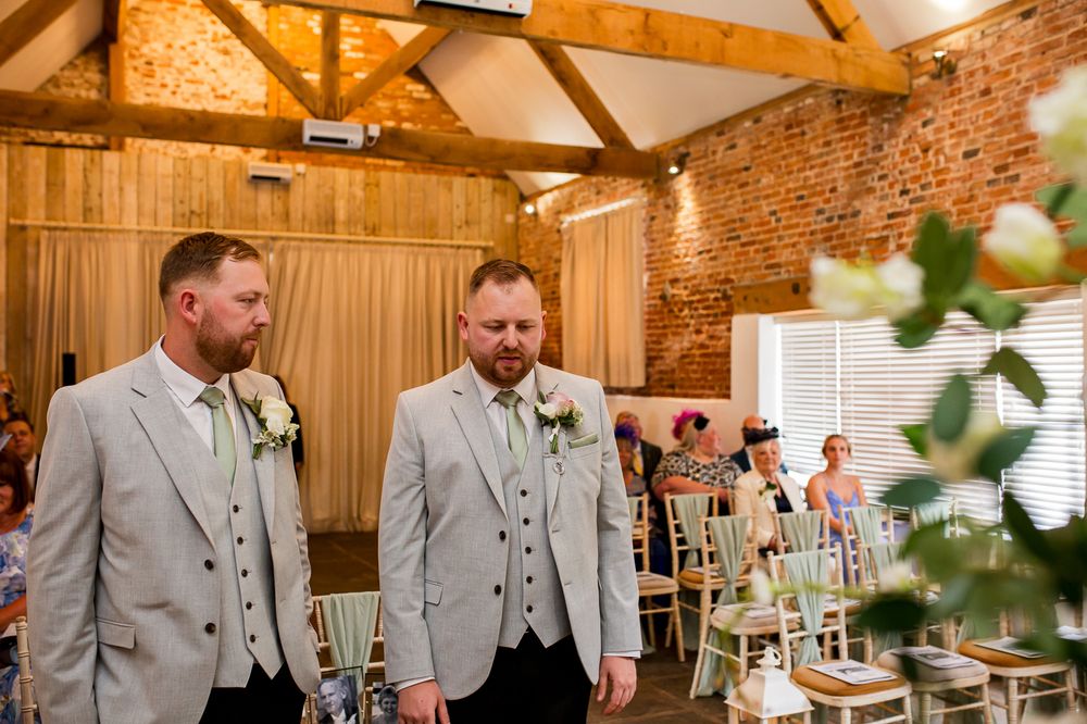 Two groomsmen in matching light gray suits stand at the front of a rustic barn wedding venue with exposed brick walls.
