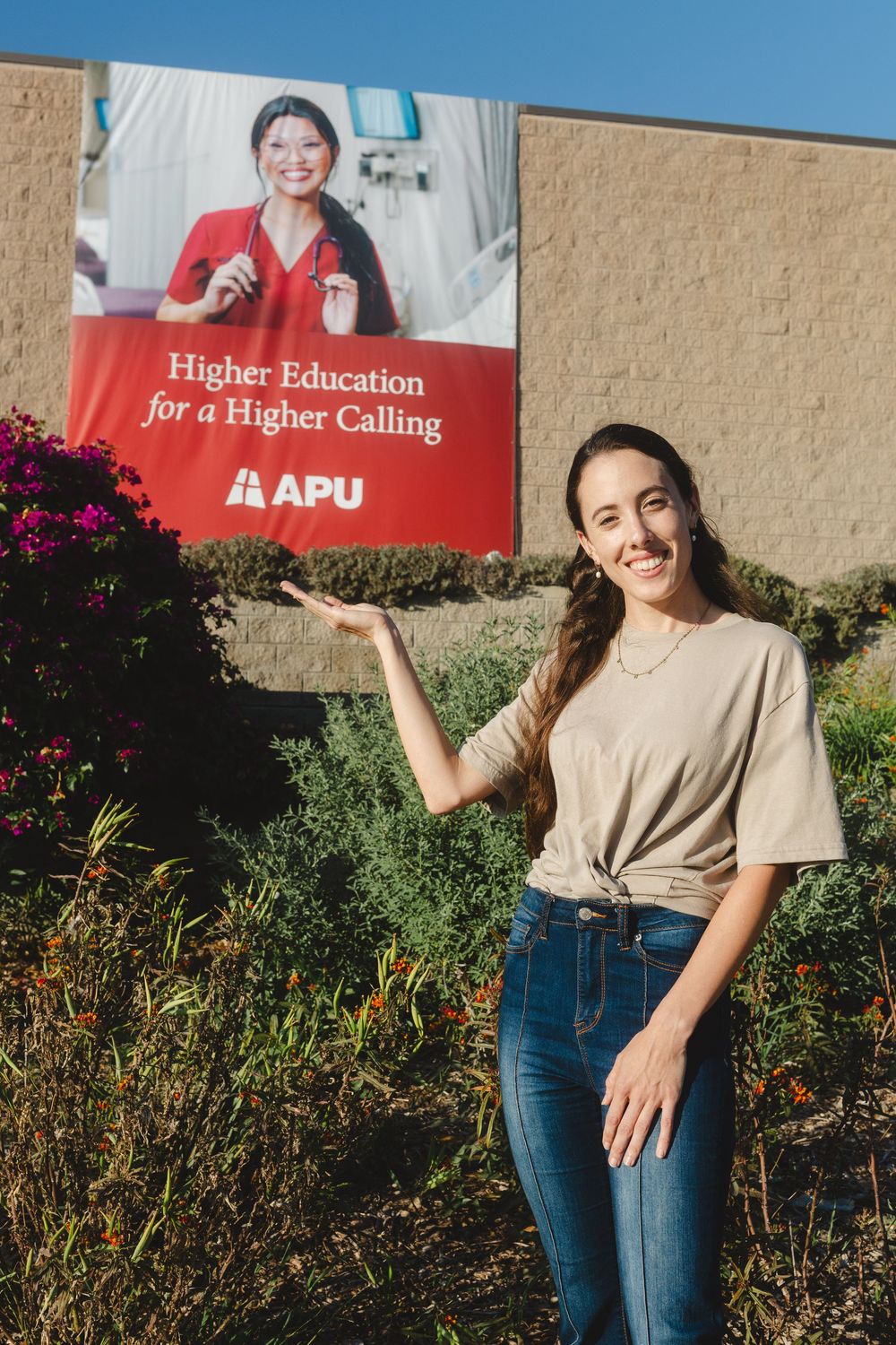 Person in casual beige top and jeans poses outdoors against garden backdrop with red banners visible.