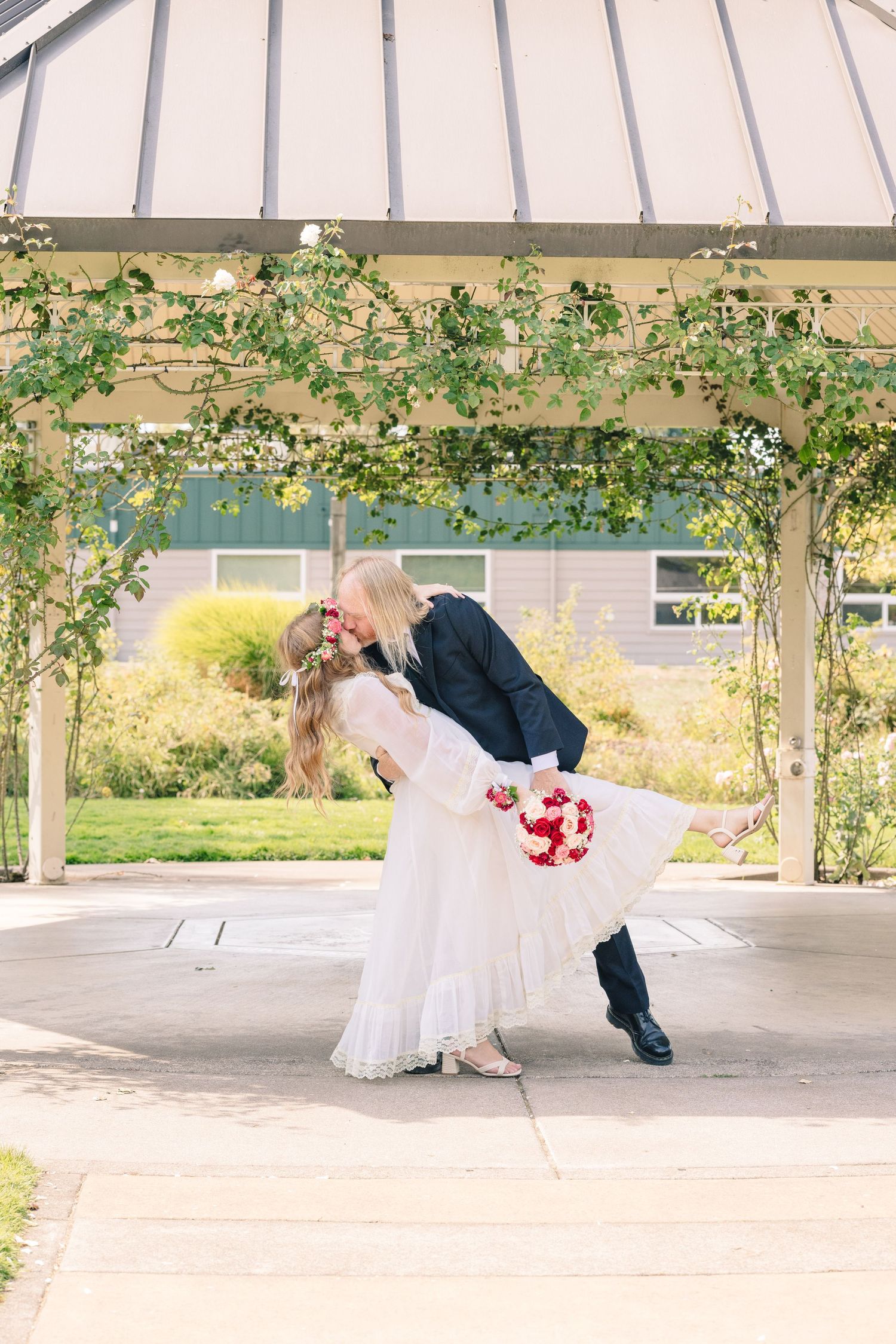 Romantic wedding couple poses under white gazebo with climbing vines and flowers.